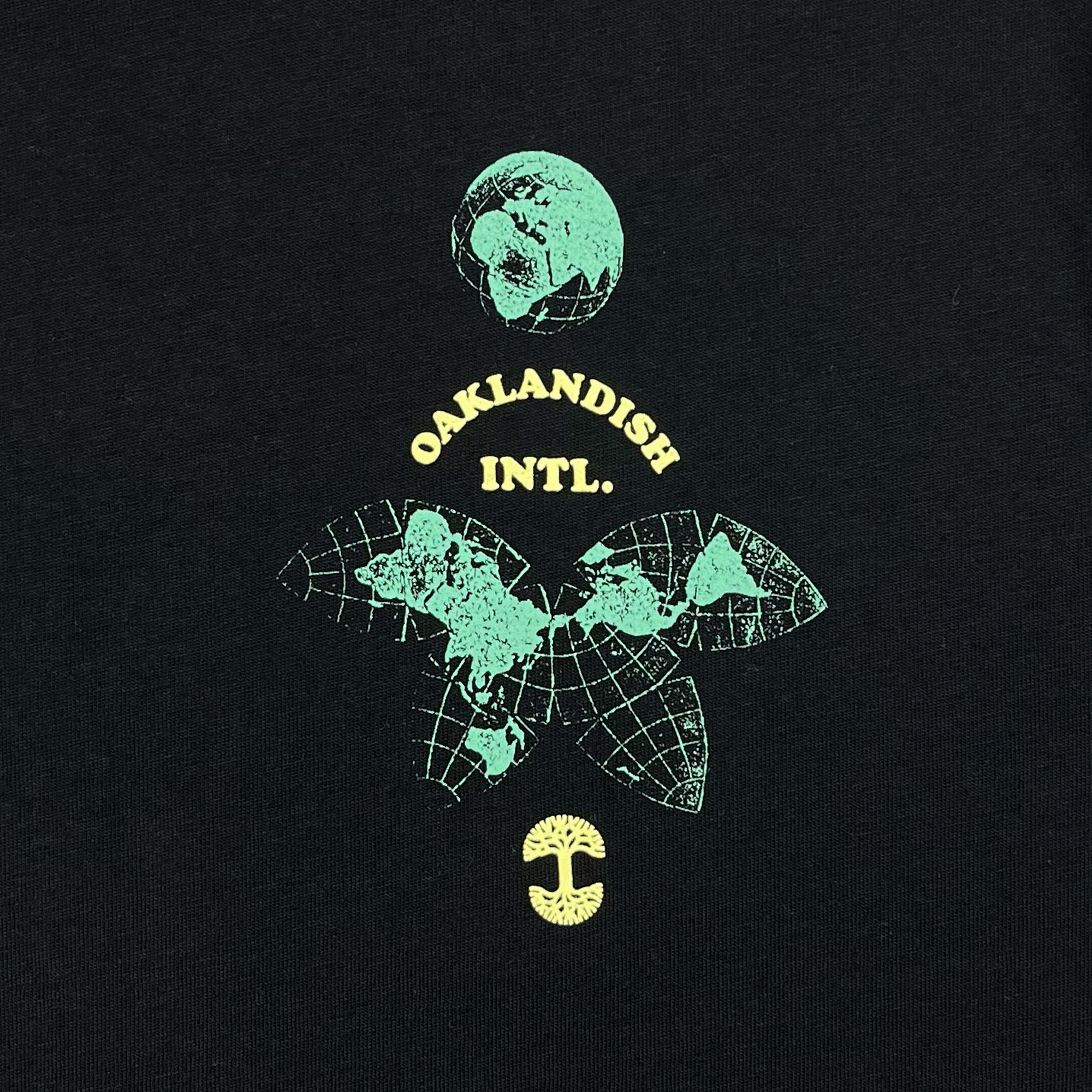 Close-up of the small Oakland International graphic and Oaklandish tree logo on the upper chest of a black t-shirt.