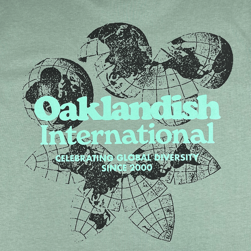 Close-up of Oakland International graphic with a caption celebrating global diversity since 2000 on a sage green tee. 