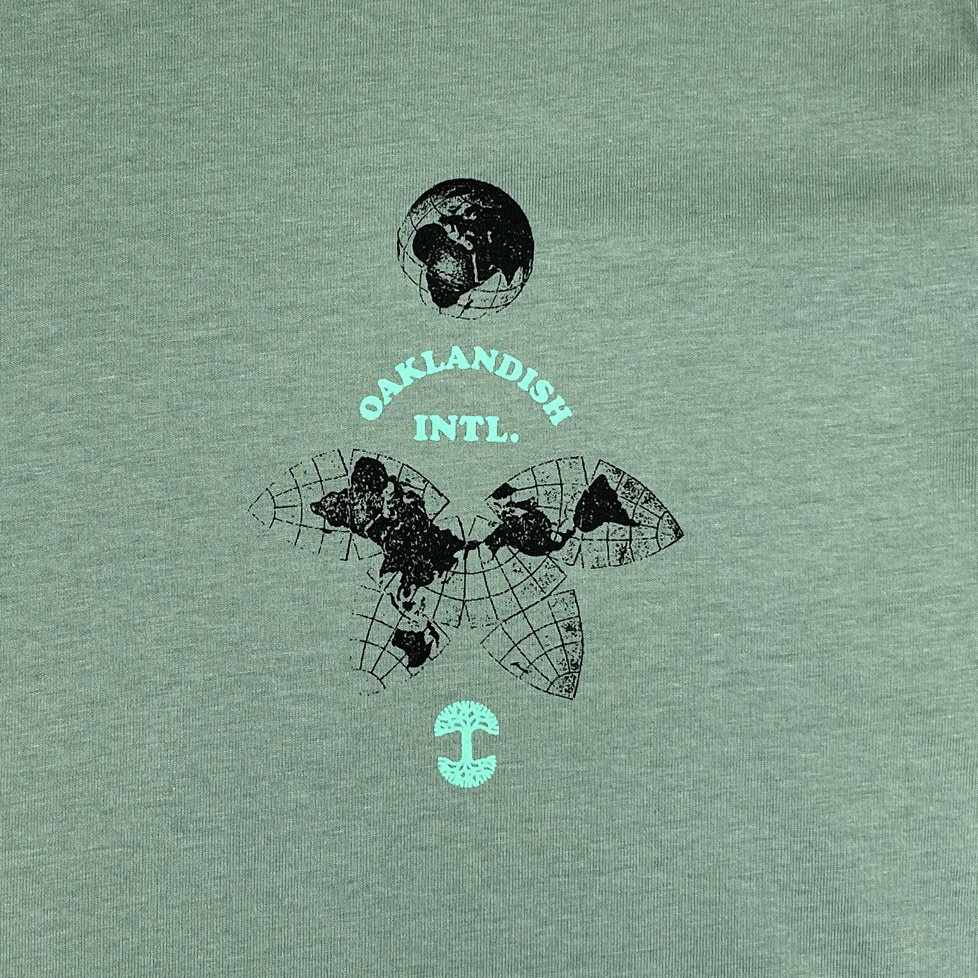 Close-up of the small Oakland International graphic and Oaklandish tree logo on the upper chest of a sage green t-shirt.