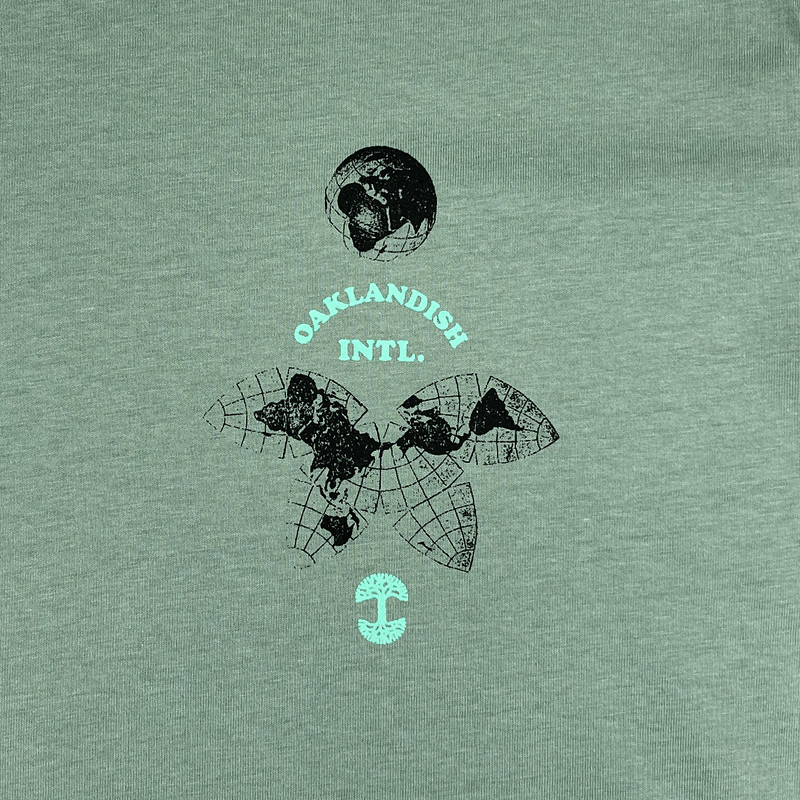 Close-up of the small Oakland International graphic and Oaklandish tree logo on the upper chest of a sage green t-shirt.
