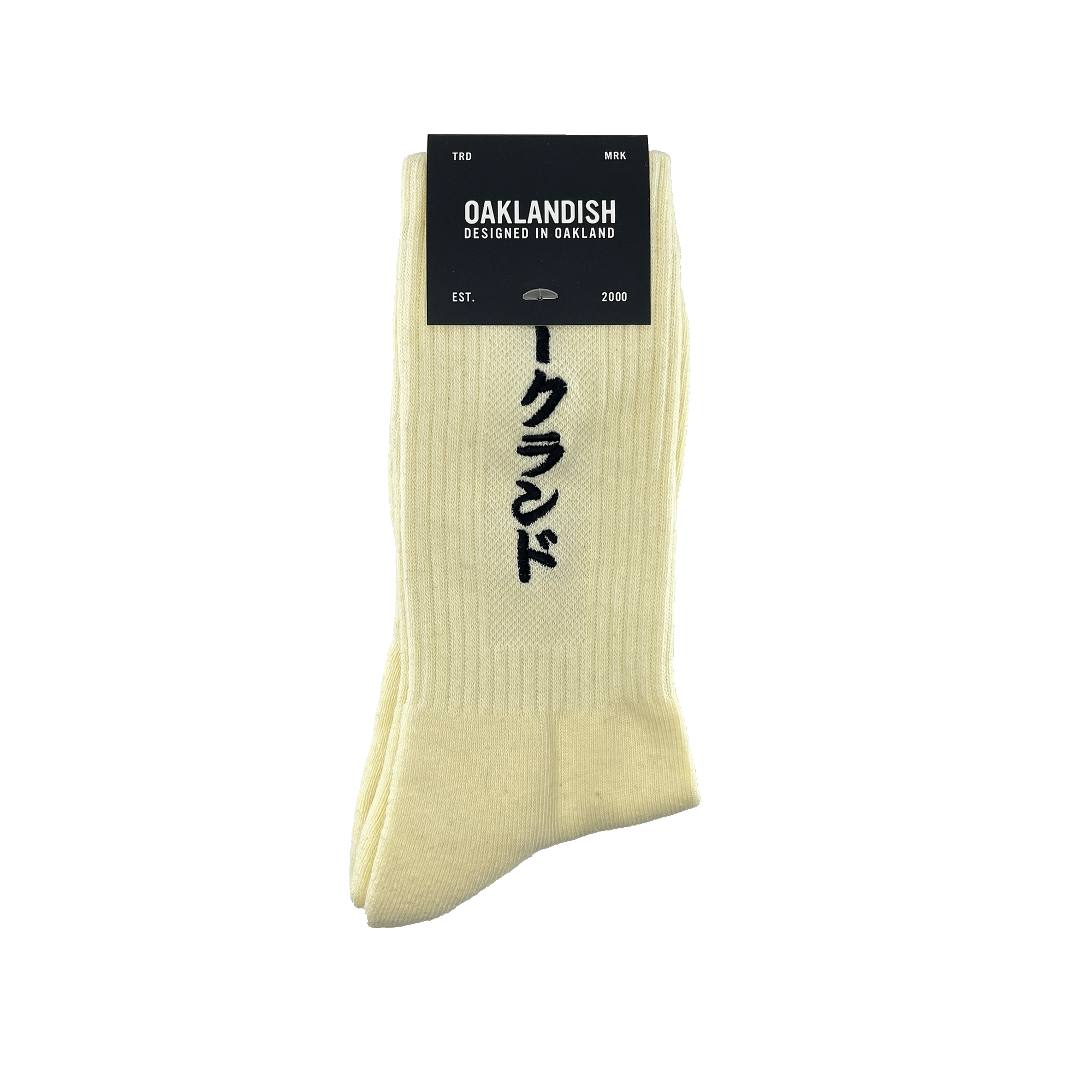 Product Description: The Kanji Sock by Oaklandish is a pair of cream-colored cotton crew socks featuring vertical Japanese characters. A black label at the top reads "OAKLANDISH" with "DESIGNED IN OAKLAND" below it. The label also includes "TRD MRK," "EST.," "2000," and a tree logo. Available in adult sizes.