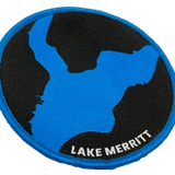 Close-up of black woven iron-on patch with a blue outline of Oakland's Lake Merritt & Lake Merritt wordmark.
