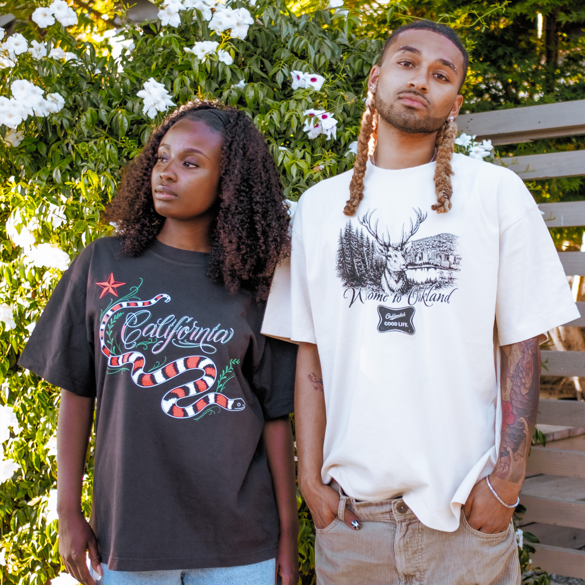 Two people stand outdoors before greenery. The left wears a black "California" snake tee, while the right sports Oaklandish's Buck Tee, a heavy-weight cotton shirt with an elk and forest illustration.