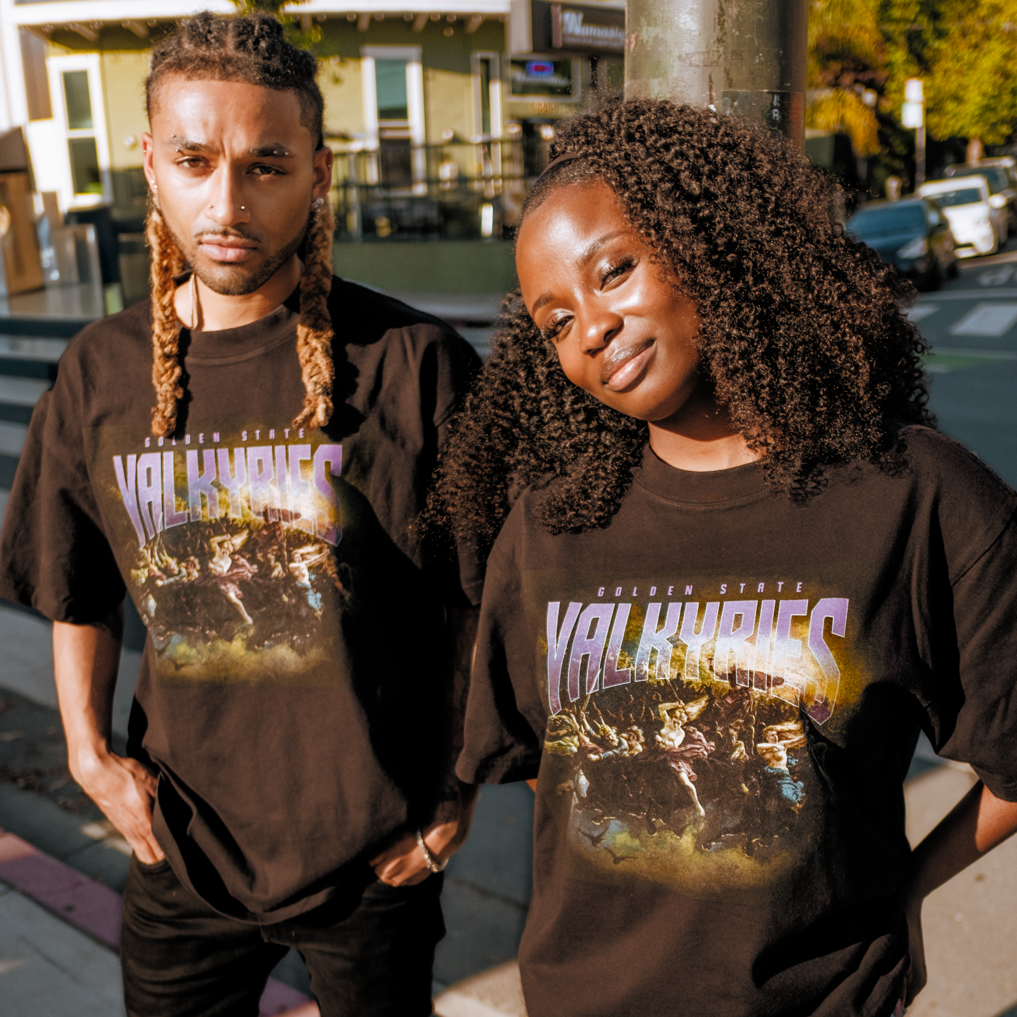 Two people stand outdoors in matching black GSV Odin Tees. The person on the left has long blond braids and a beard; the right, with curly hair, smiles as sunlight highlights their 100% cotton shirts against a city backdrop.