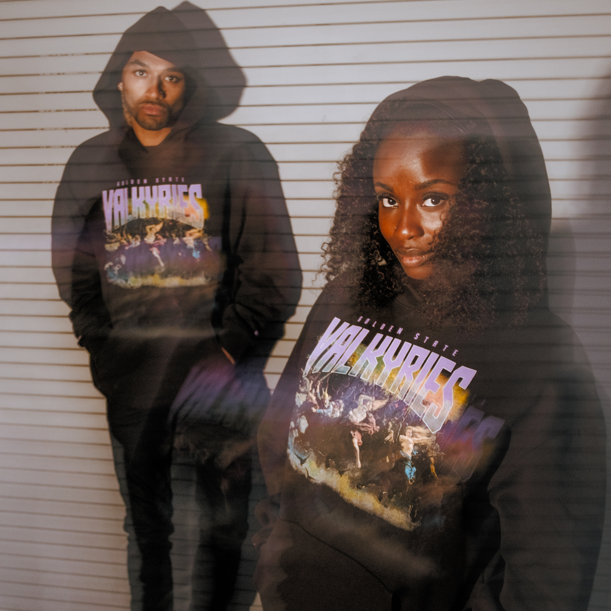 Two people pose against a white backdrop, both wearing GSV Odin Hoodies in black with colorful graphics. The woman faces forward; the man stands in shadow behind her. The photo features a soft, double-exposure effect.