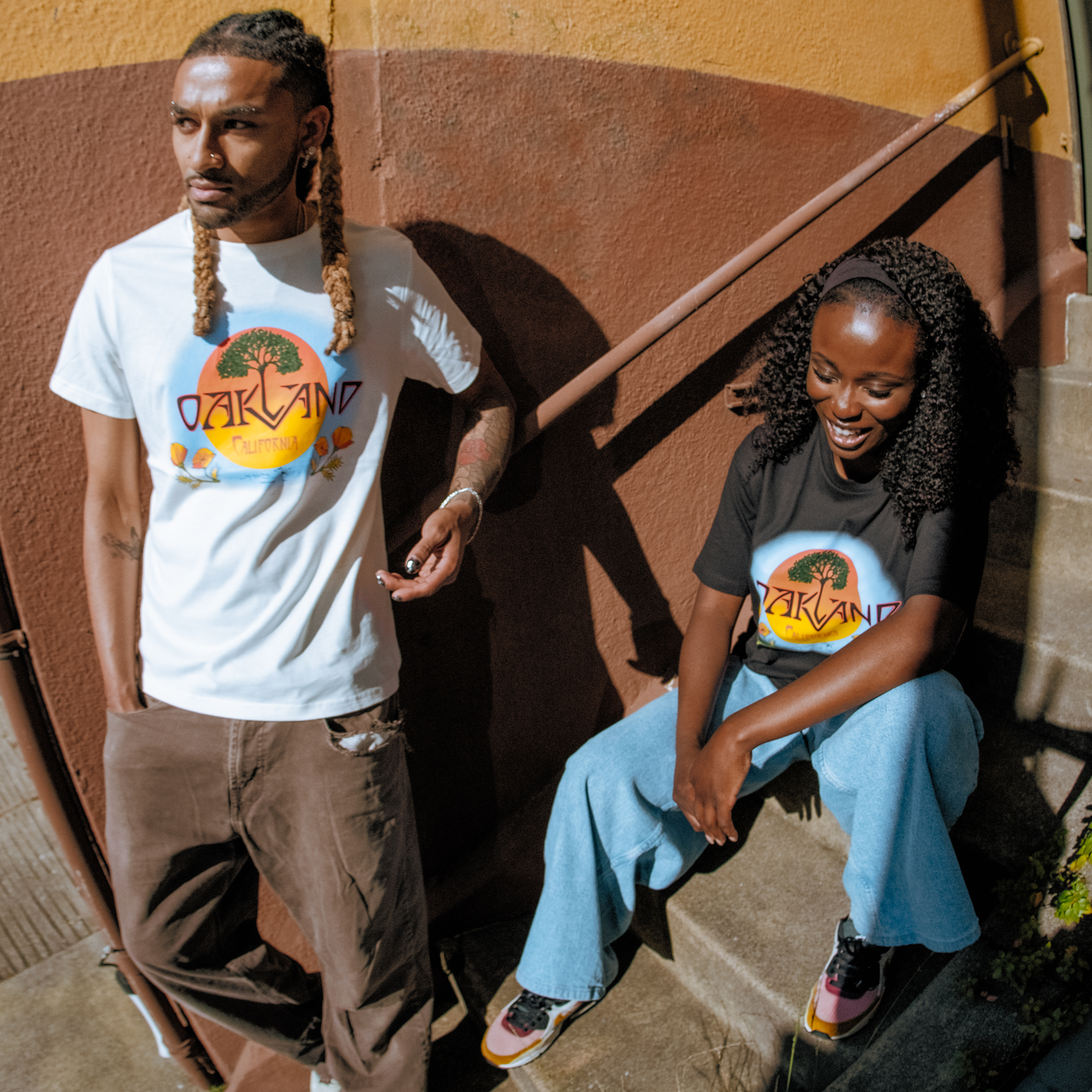 Two people wear Oaklandish Vanguard Tees with “Oakland, California” graphics as they relax on concrete steps by a mustard-yellow and brown wall. The man stands casually while the woman smiles; both pair their shirts with jeans and sneakers in the sunlight.