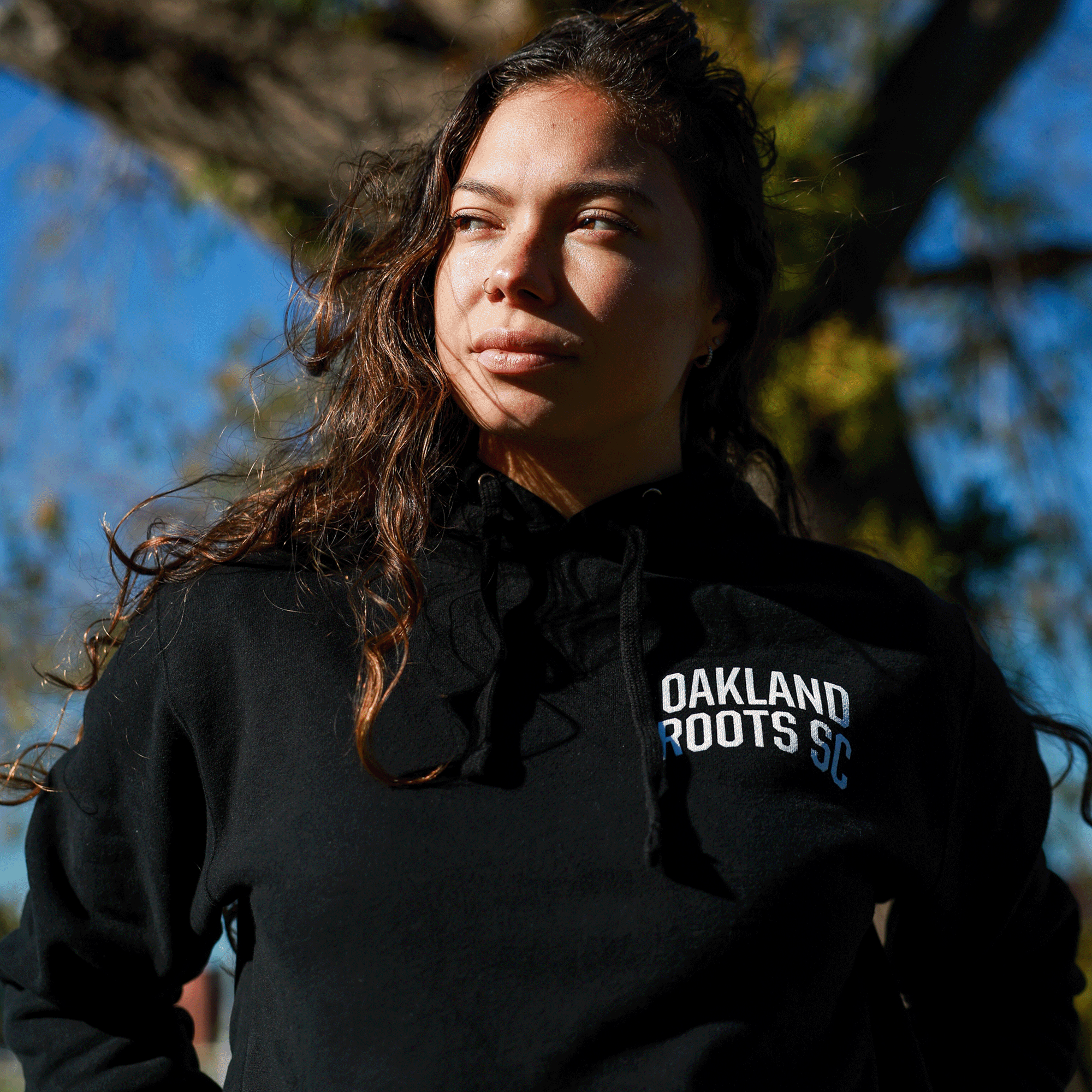 Model wearing black pullover hoodie with white OAKLAND ROOTS SC wordmark on the chest.