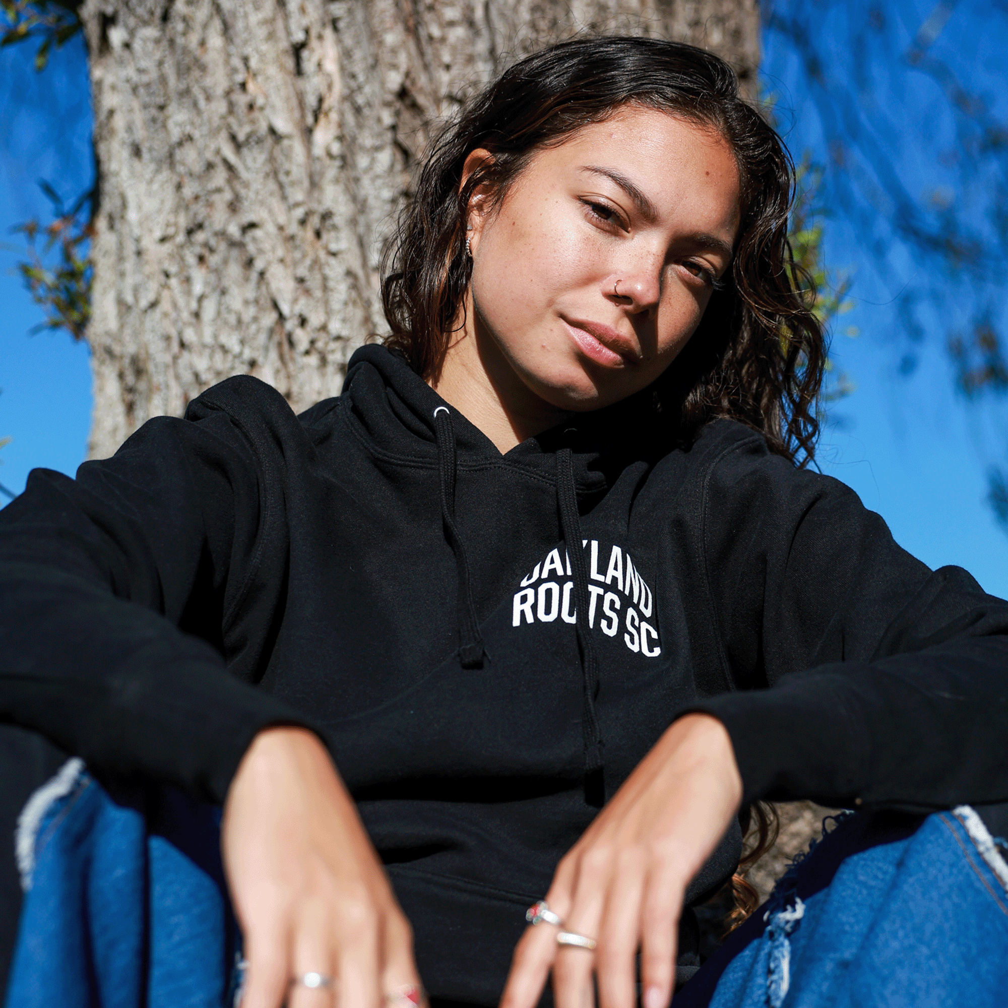 Model wearing black pullover hoodie with white OAKLAND ROOTS SC wordmark on the chest.