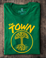 green shirt folded on a piece of wood, shirt has a design in golden yellow ink saying the town and then the Oaklandish tree with roots logo inside of a circle.