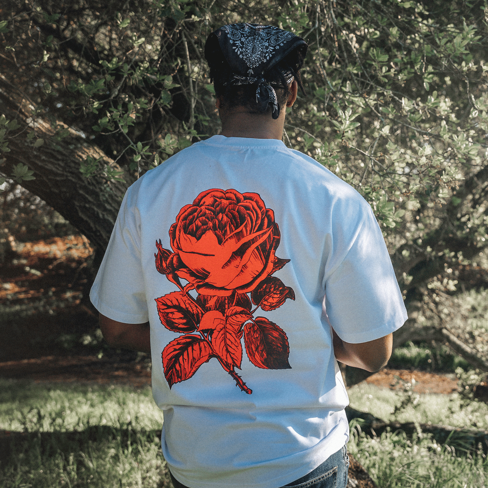 A person wearing a blue bandana and an Oaklandish Town Rose Heavy Tee, characterized by its boxy fit and heavyweight combed cotton material, stands outdoors with their back to the camera. The T-shirt features a large red rose design on the back. The background is filled with lush green trees and dappled sunlight, suggesting a serene, natural setting.