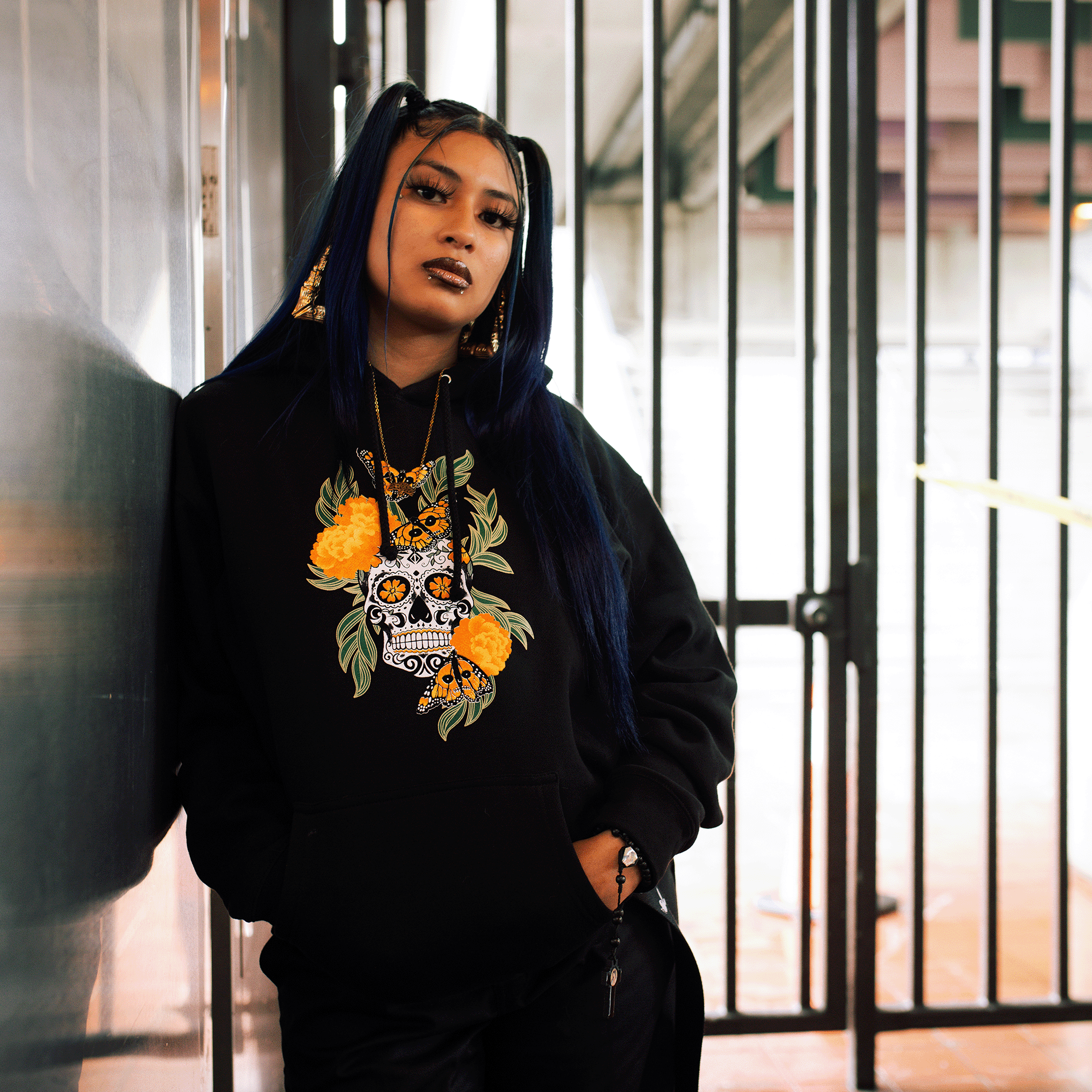 Female model wearing black pullover hoodie with graphic art by Oakland artist Jet Martinez depicting a sugar skull surrounded by Marigolds and Monarch butterflies.