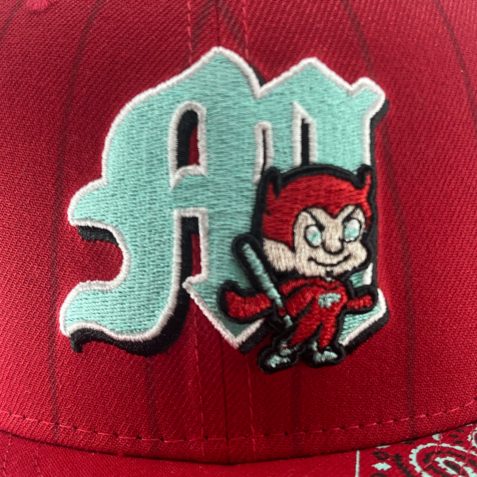 A close-up shot of the New Era Diablos Pinstripe 59FIFTY Cap from Retail Vendors, showcasing a detailed embroidered logo. The red cap features stylized green letters "AM" and a small cartoon devil character holding a baseball bat, positioned to the right of the letters. The devil has red skin, horns, and a mischievous smile on this 100% polyester cap.