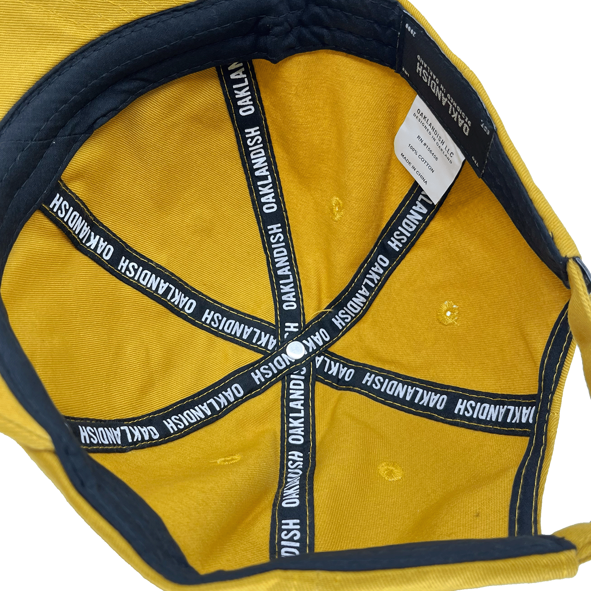 Interior view of the yellow Micro Logo Dad Hat by Oaklandish, showcasing navy blue fabric lining with "OAKLANDISH" in white text repeatedly along the seams. The cap features an adjustable strap back and a white tag with washing instructions sewn into the inner edge.
