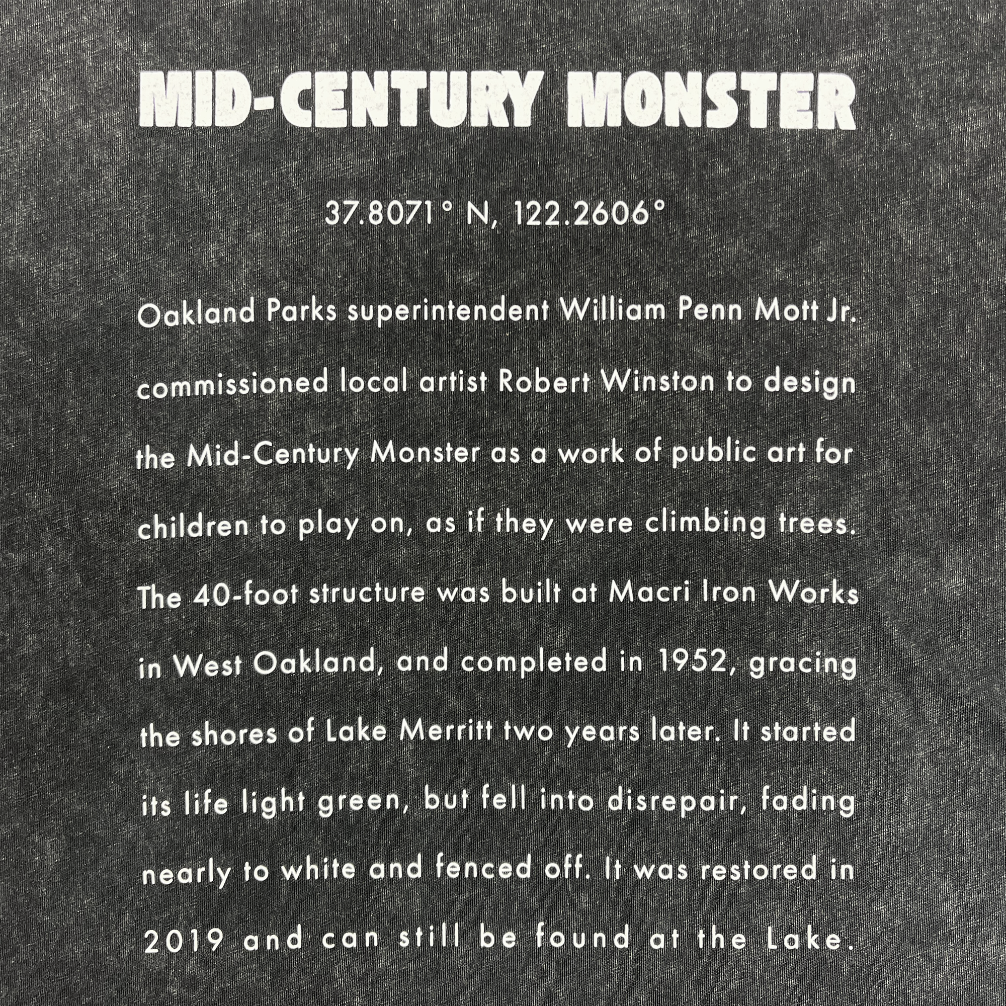 Detailed close-up long-form text explaining the history of the mid-century monster structure at Oaklands Marci Iron Works on the back of a black stone-colored t-shirt.