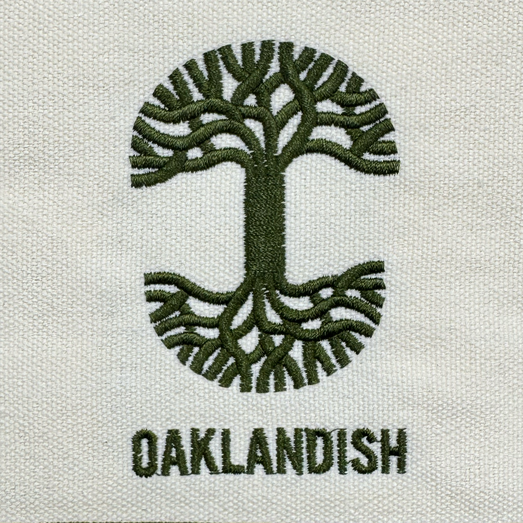 The Oaklandish Mini Grocery Tote features a green embroidered tree with spreading branches and roots forming an oval. Below it, "OAKLANDISH" is stitched in green capitals on textured beige fabric. Brand: Oaklandish.