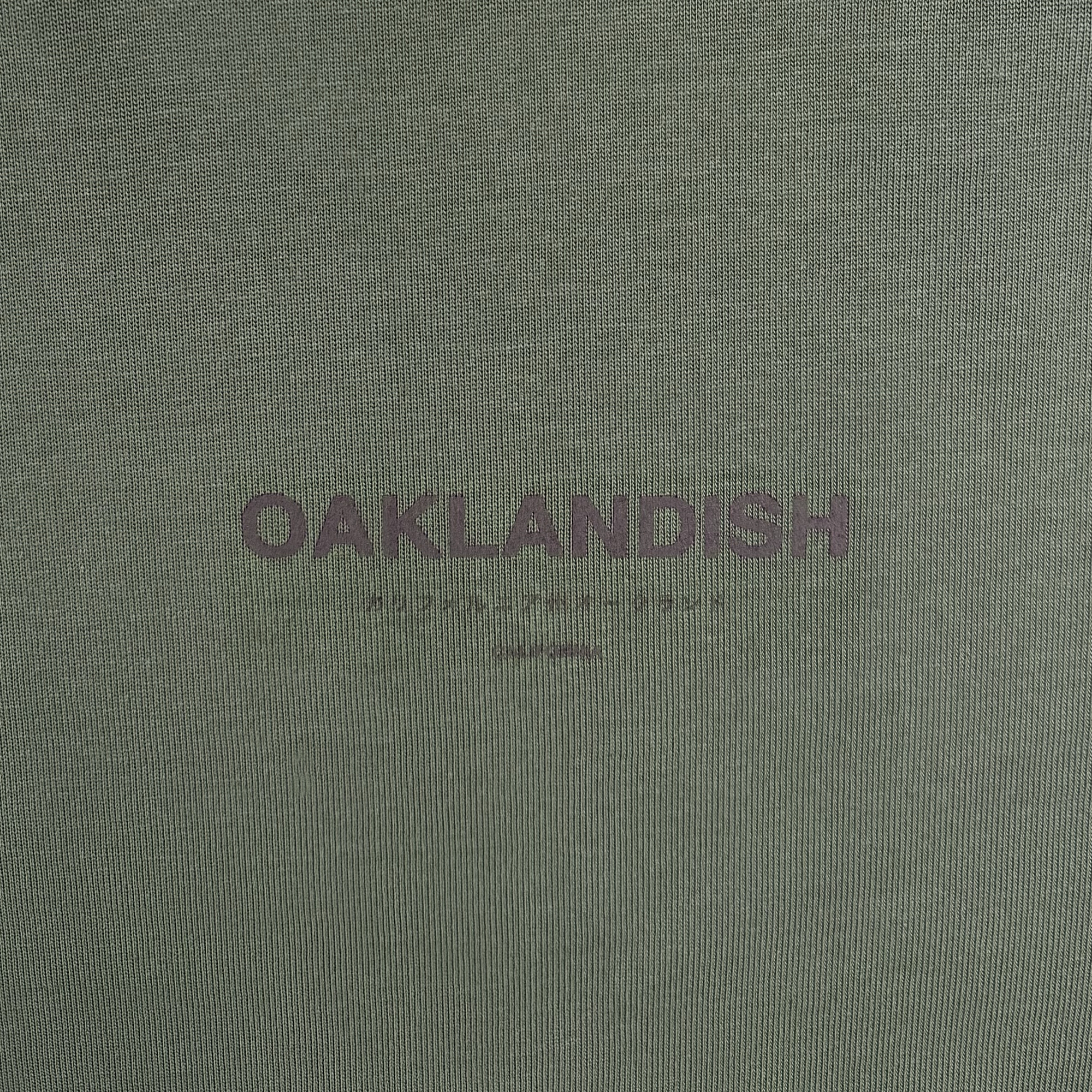 Close-up of the Modern Type Heavy Tee by Oaklandish, featuring a heavyweight, boxy fit with green fabric. The shirt showcases "OAKLANDISH" in bold, dark lettering at the center, accompanied by "the Higher Spirit" and "OAKLAND" in smaller text below. The texture of the 100% carded cotton fabric is clearly visible.