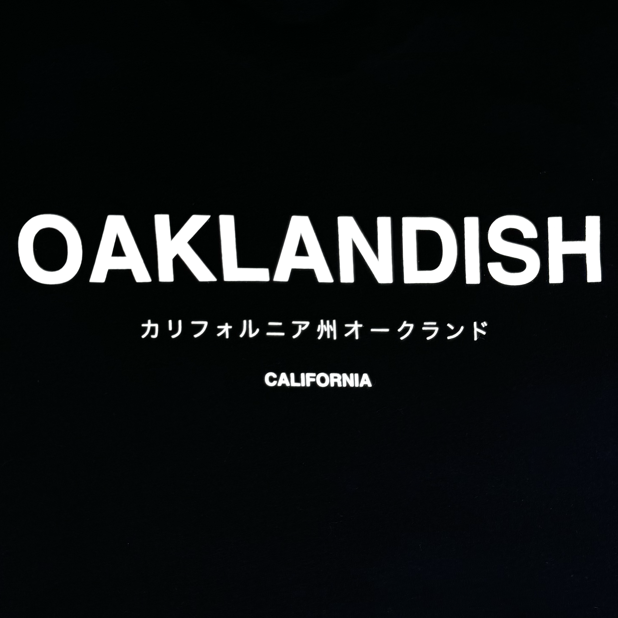 The Modern Type Tee by Oaklandish features a black background with bold white "OAKLANDISH" text at the top. Below, Japanese characters read "カリフォルニア州オークランド," translating to "Oakland, California," and further down, smaller white text spells out "CALIFORNIA." This design is available on a comfortable cotton t-shirt for men.