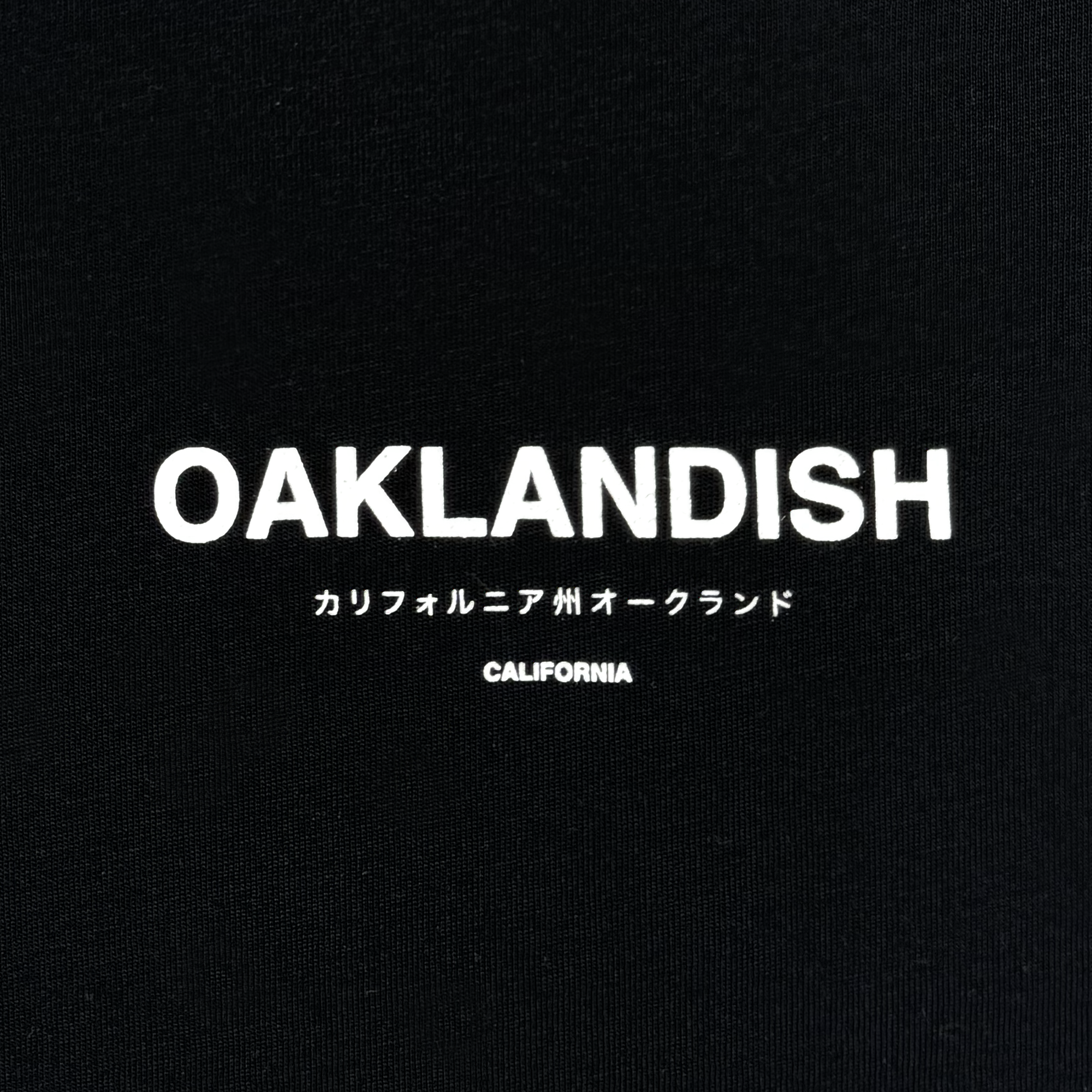 The image showcases a black background with the word "OAKLANDISH" prominently displayed in bold white letters, ideal for the Oaklandish Modern Type Tee crafted from men's cotton. Below, Japanese characters translate to "California Oakland," with "CALIFORNIA" subtly printed in small white text beneath.