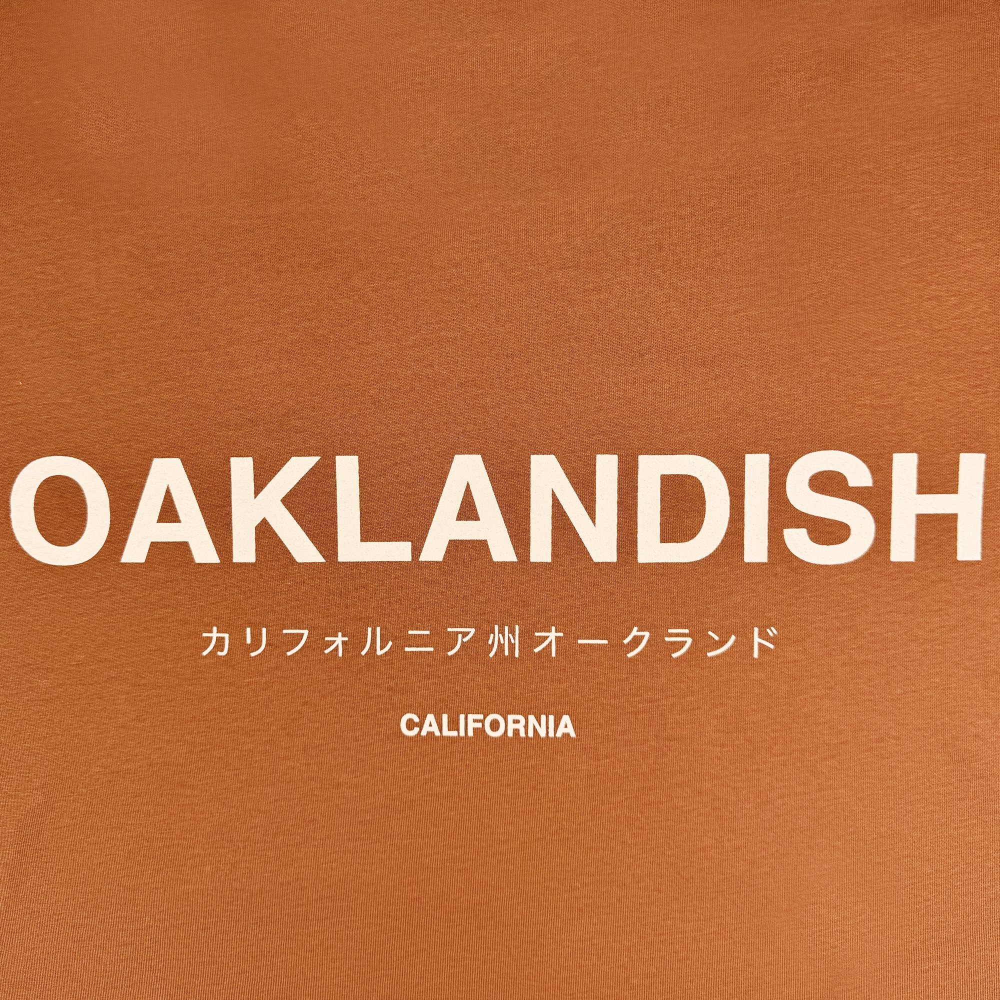 The image showcases the "Modern Type Tee" by Oaklandish, featuring "OAKLANDISH" in bold white letters on a brown t-shirt. Below, Japanese characters spell "カリフォルニア州オークランド," with "CALIFORNIA" centered at the bottom. This sleek design is perfect for men, crafted from 100% cotton for ultimate comfort.