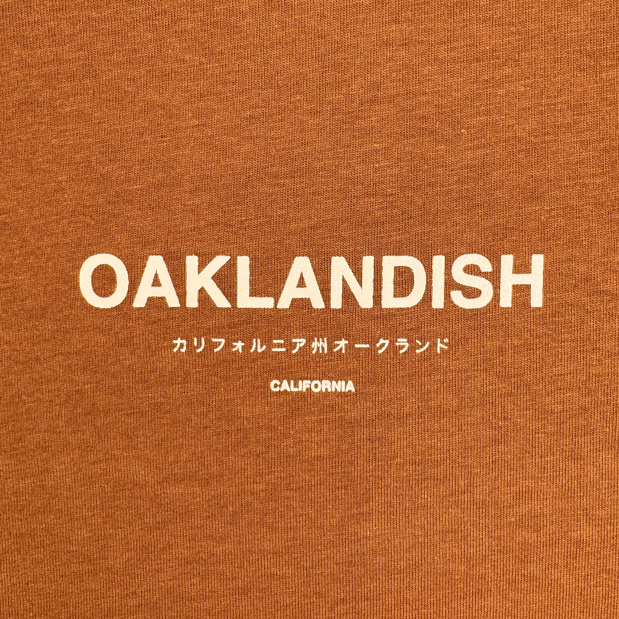 The image showcases the text "OAKLANDISH" in bold white font on a brown background, similar to a stylish t-shirt design. Below are Japanese characters that translate to "California, Oakland," with "CALIFORNIA" in smaller white font. The Modern Type Tee by Oaklandish is perfect for men looking for a 100% cotton blend of style and comfort.
