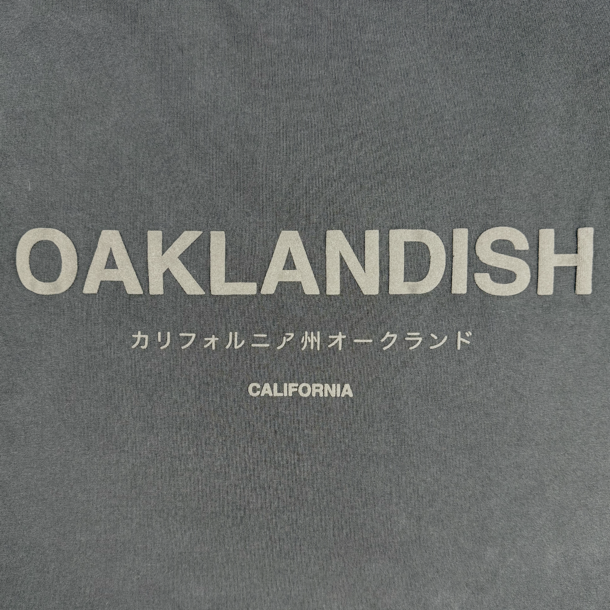 The Modern Type Heavy Tee by Oaklandish is a heavyweight cotton shirt in faded black, featuring "OAKLANDISH" in bold light gray text centered on the front. Below, it includes Japanese text and the word "CALIFORNIA" in a smaller matching font.