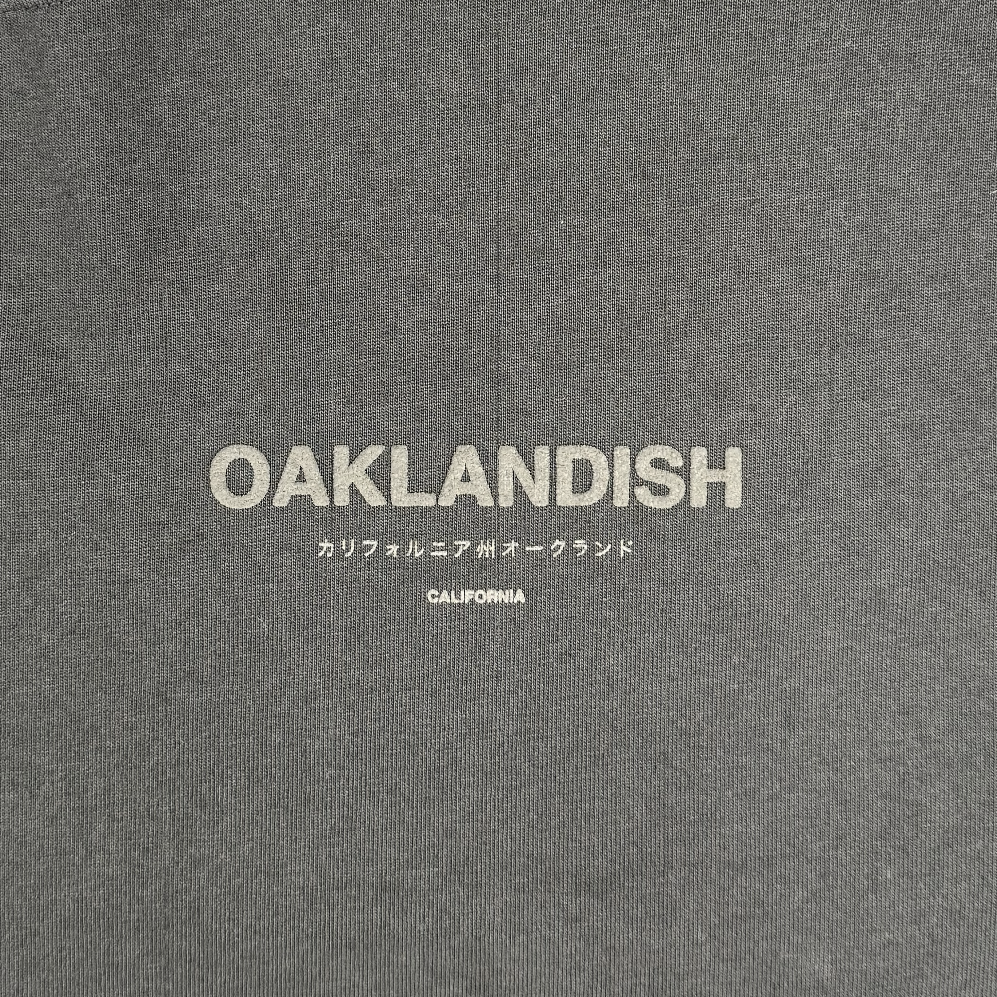 The Modern Type Heavy Tee from Oaklandish features a faded black fabric with "OAKLANDISH" in bold white letters, complemented by Japanese characters and "CALIFORNIA" beneath them in smaller white lettering. The texture suggests it's made of heavyweight cotton.