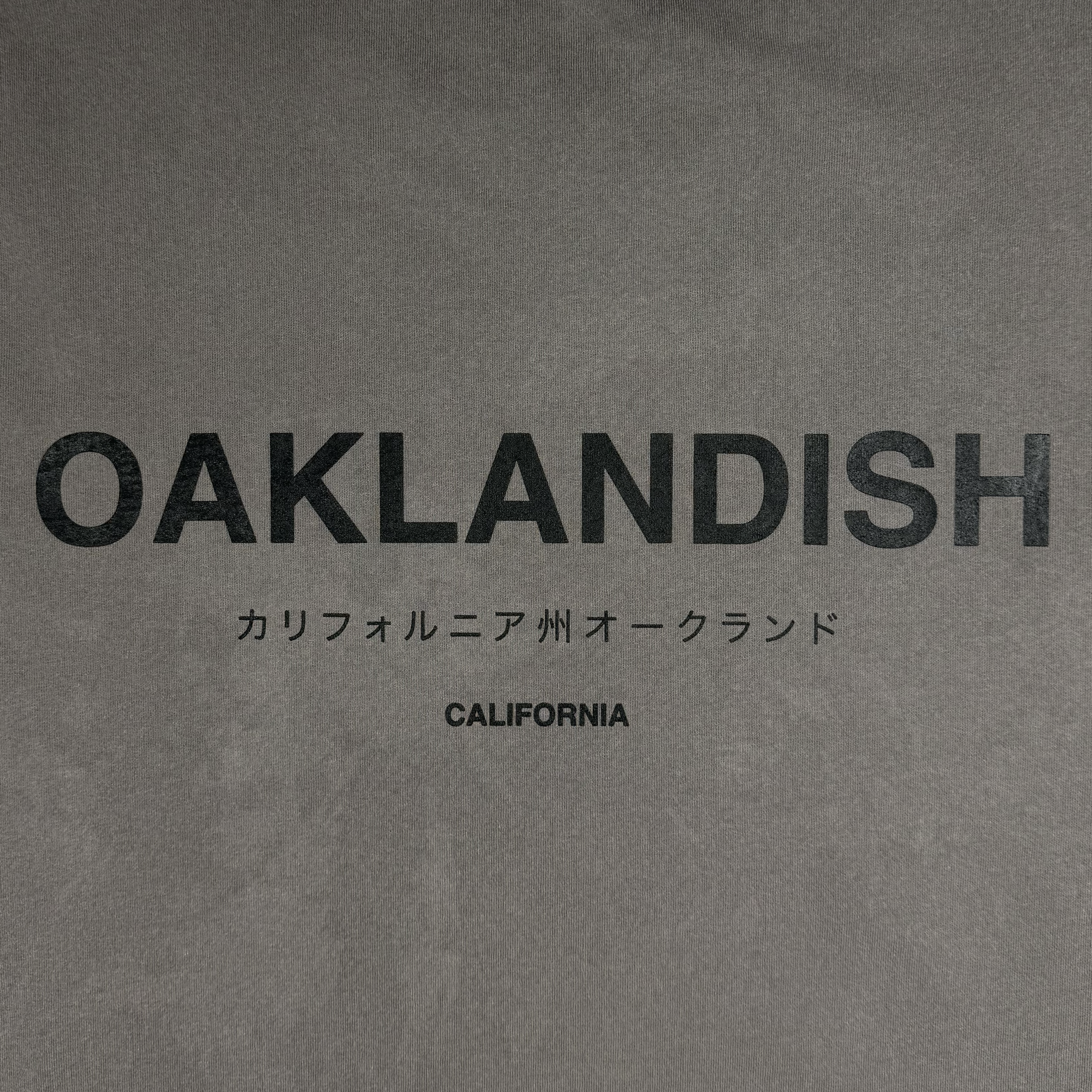 The Modern Type Heavy Tee by Oaklandish features a gray background with "OAKLANDISH" in bold black letters at the top. Below, Japanese characters translate to "California Oakland," and the word "CALIFORNIA" is printed in smaller black text at the bottom. Made from 100% carded cotton, this heavyweight tee provides a classic look and feel.