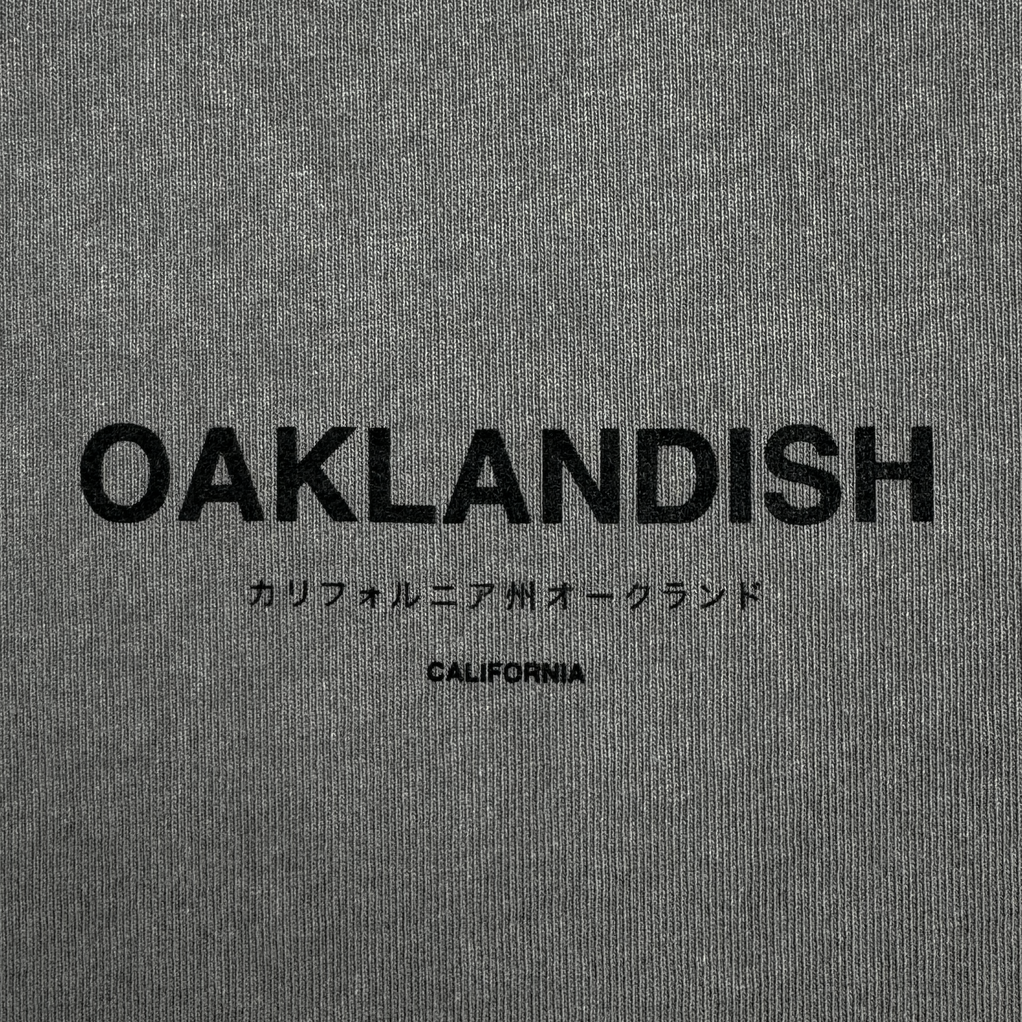The Modern Type Heavy Tee by Oaklandish showcases a gray fabric with "OAKLANDISH" prominently displayed in bold black letters. Below it, Japanese characters translate to "California Oakland," and the word "CALIFORNIA" appears underneath in a smaller font. Crafted from 100% carded cotton, its boxy fit provides both comfort and style.