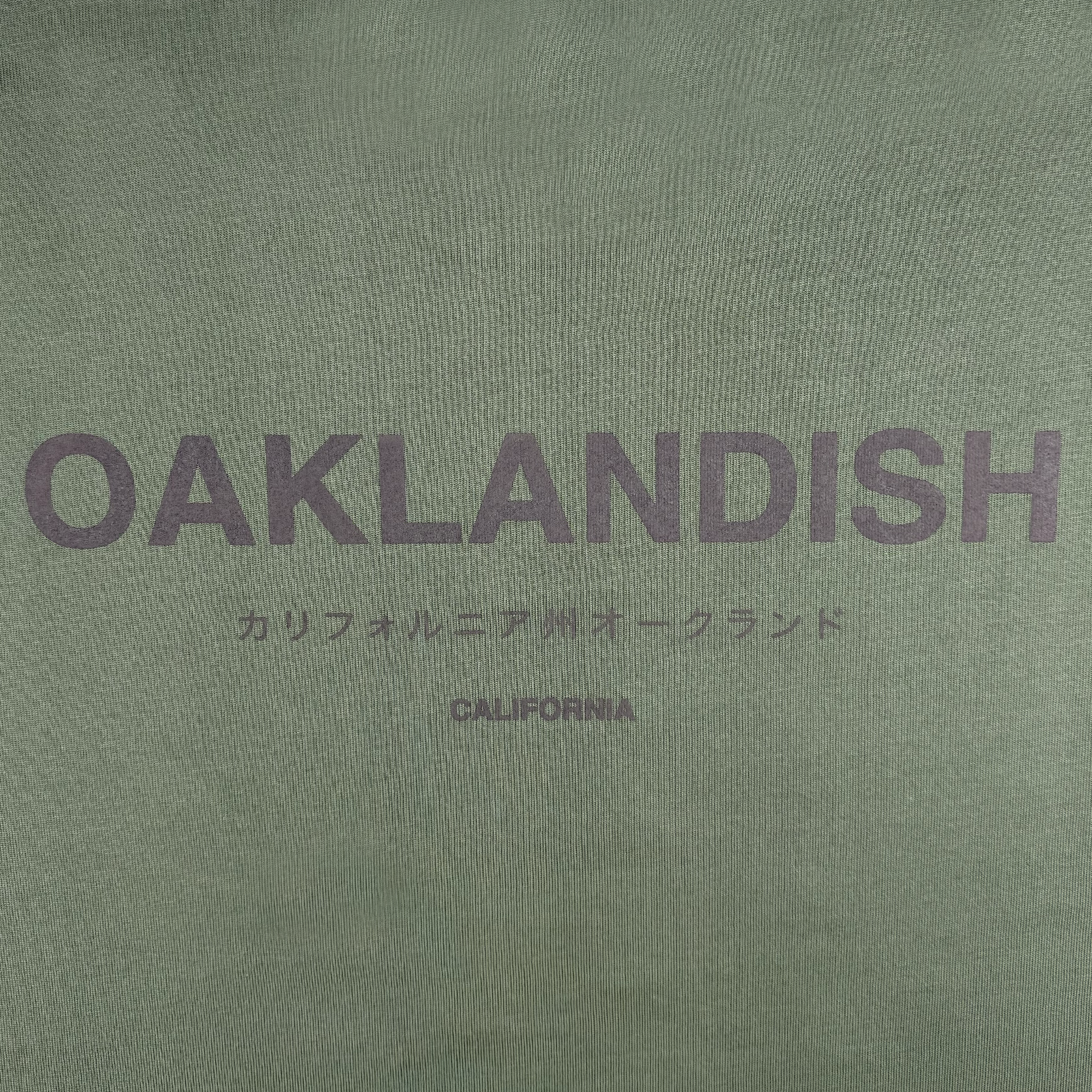 The Modern Type Heavy Tee by Oaklandish features a green heavyweight textured fabric with "OAKLANDISH" prominently printed in bold, dark letters. Underneath, Japanese characters can be seen along with "CALIFORNIA" in smaller font. Made from 100% carded cotton, this t-shirt boasts a boxy fit for optimal comfort and style.