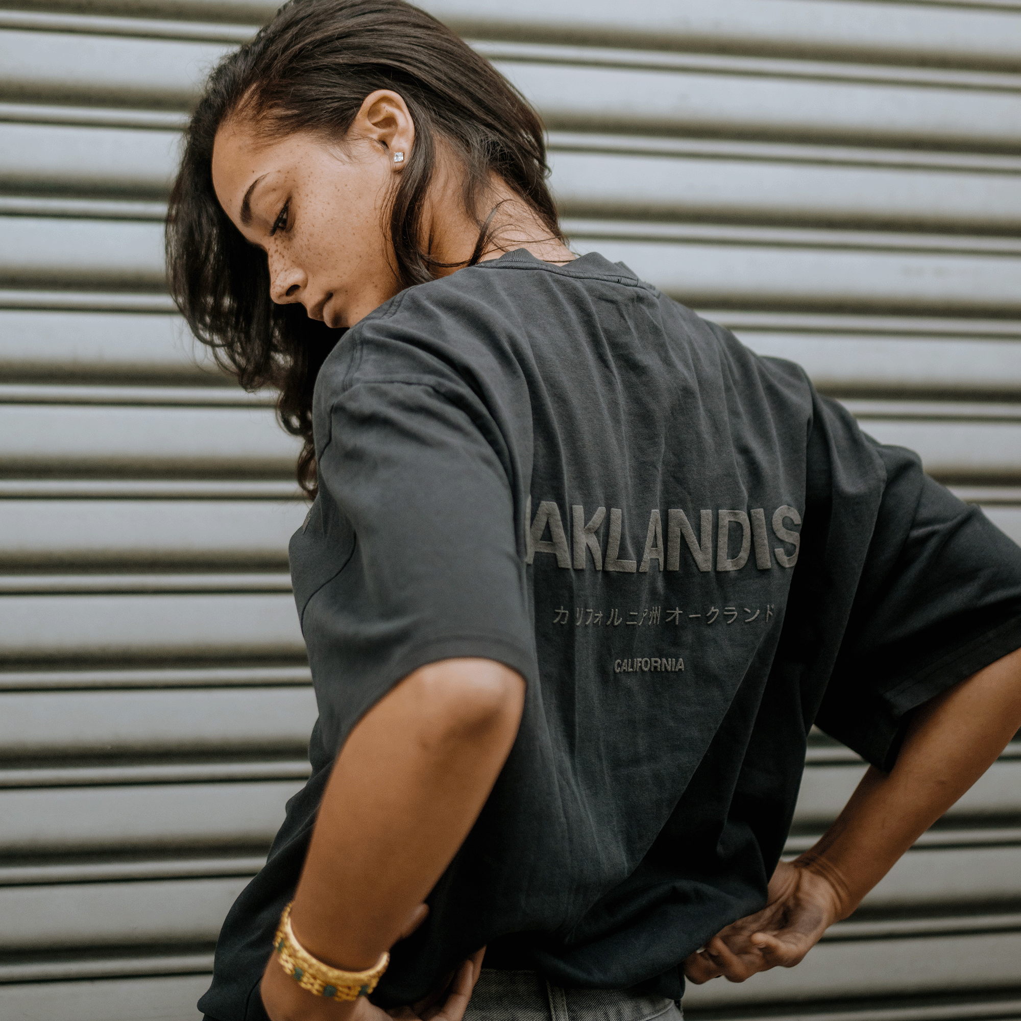 A woman with long dark hair stands calmly in front of a metal shutter, showcasing the Oaklandish Modern Type Heavy Tee, a faded black boxy fit T-shirt made from heavyweight cotton. The shirt displays the brand's name "OAKLANDISH." She wears small stud earrings and golden bracelets, posing with one hand on her hip.