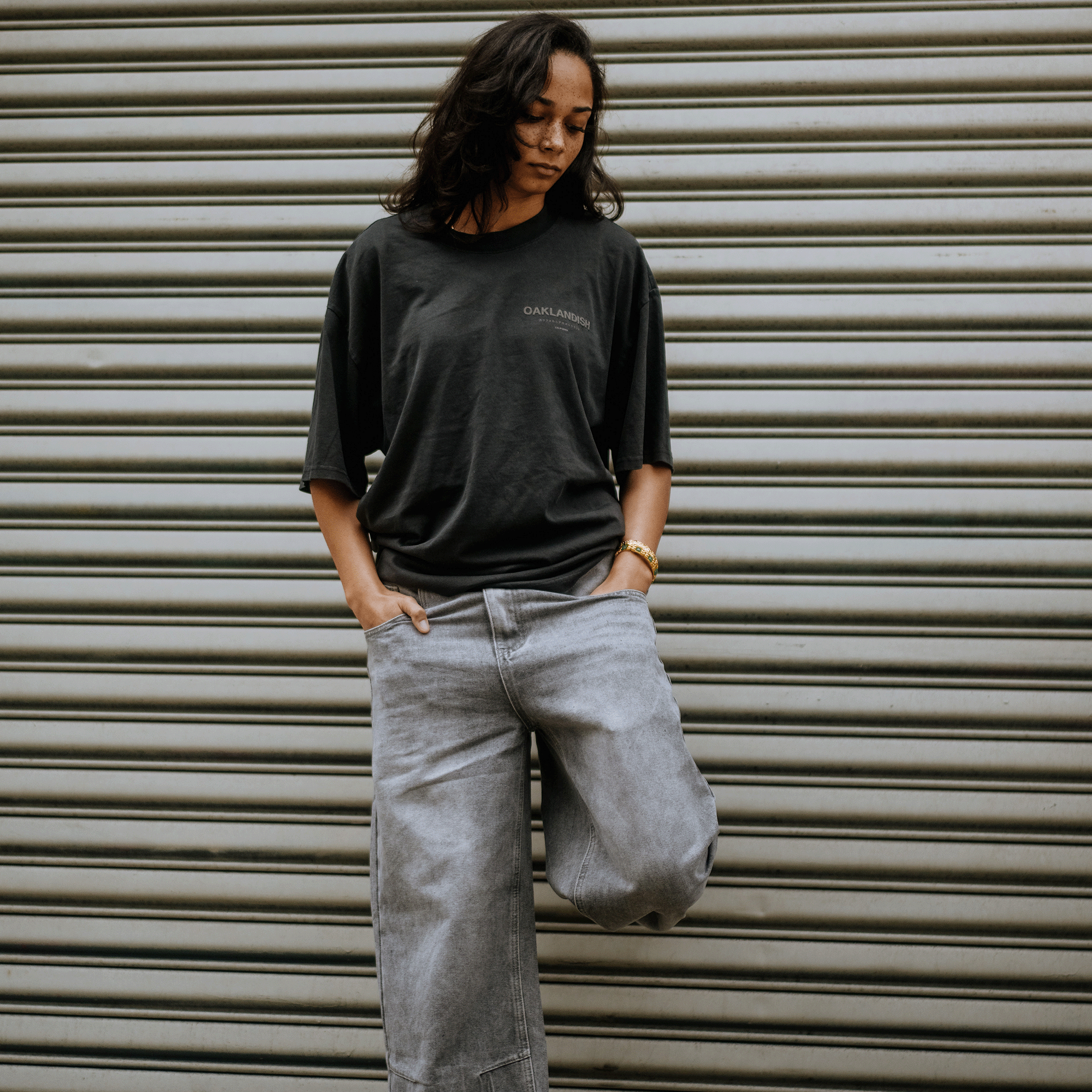 Against a metal roll-up door, a person wears the Oaklandish Modern Type Heavy Tee—a faded black, boxy fit with "Oakland" on it—and light gray jeans. Their hands are in pockets, medium-length dark hair frames their calm expression as they look downwards, suggesting an urban vibe.