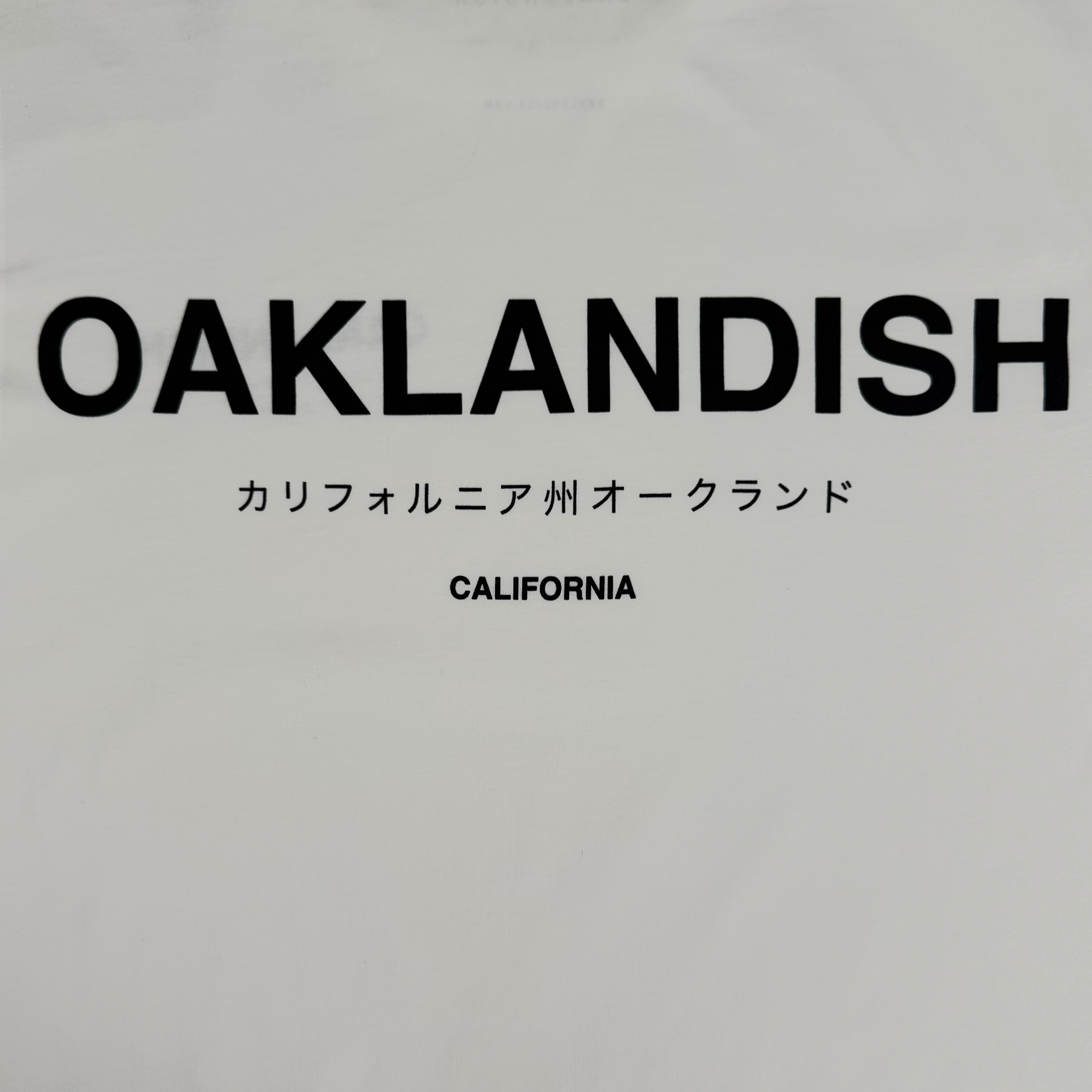 The Oaklandish Modern Type Tee features bold black "OAKLANDISH" text, Japanese script, and "CALIFORNIA" below on a white 100% cotton t-shirt. This minimalist men’s collection design has a centered layout for a modern look.