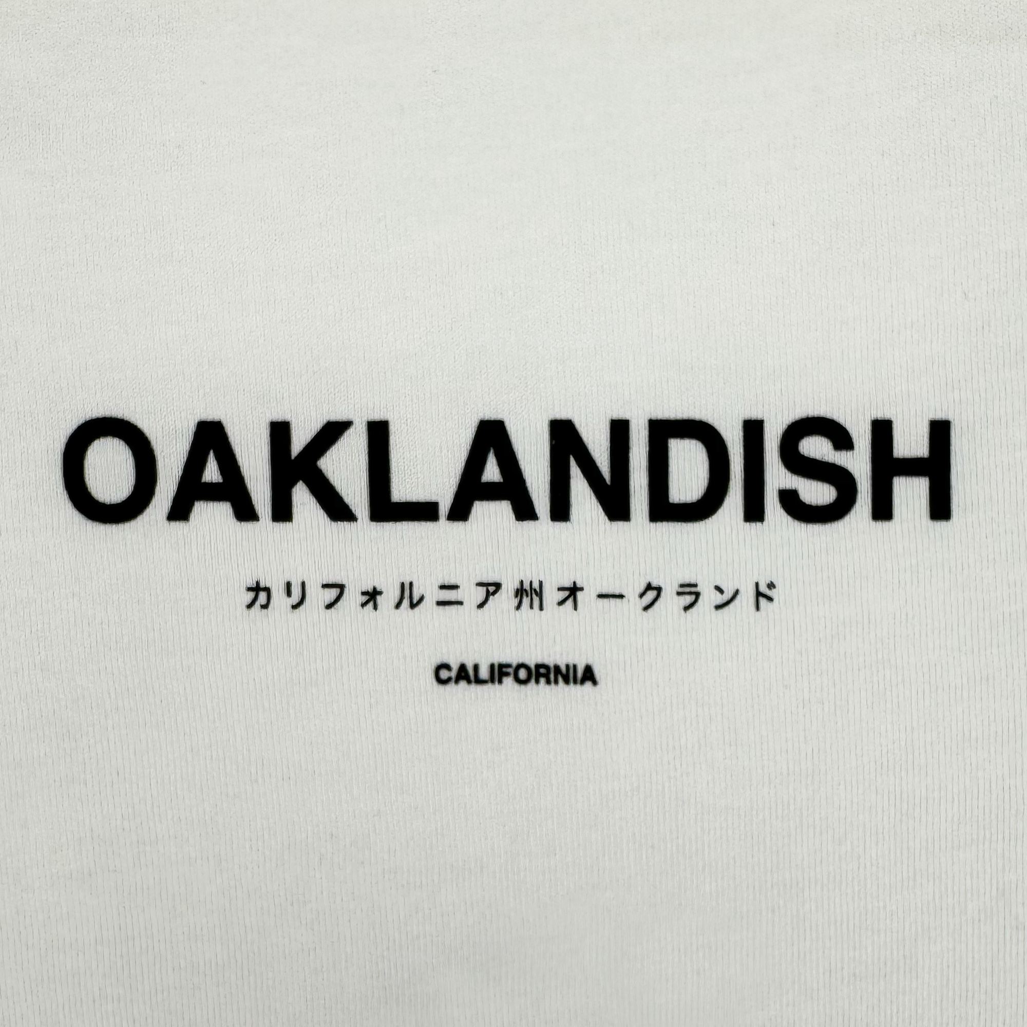 The Oaklandish Modern Type Tee features a white background with bold "OAKLANDISH," Japanese text reading "State of California, Oakland," and "CALIFORNIA" below. Made from 100% cotton and part of our men's collection.