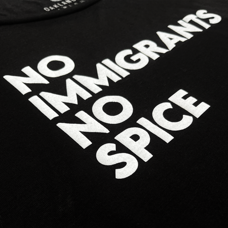 Detailed close-up of NO IMMIGRANTS NO SPICE wordmark on the front chest of a women’s black cotton tank top.