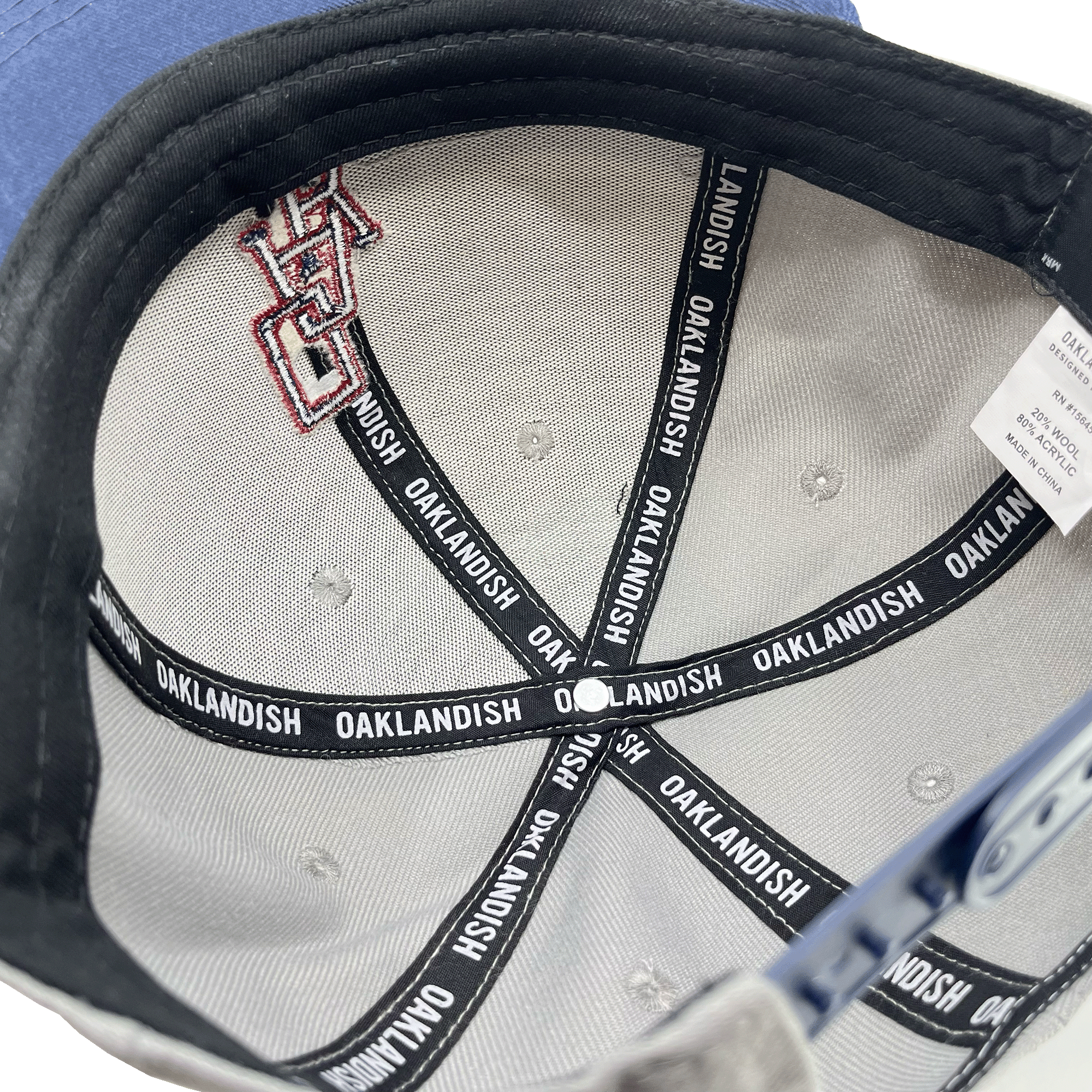 The inside of the OAK Monogram Snapback is shown, featuring light gray fabric with black reinforcing tapes. The tapes read "OAKLANDISH" repeatedly in white text. There is a label with washing instructions and size information attached. Additionally, part of an embroidered OAK logo is visible on the fabric.