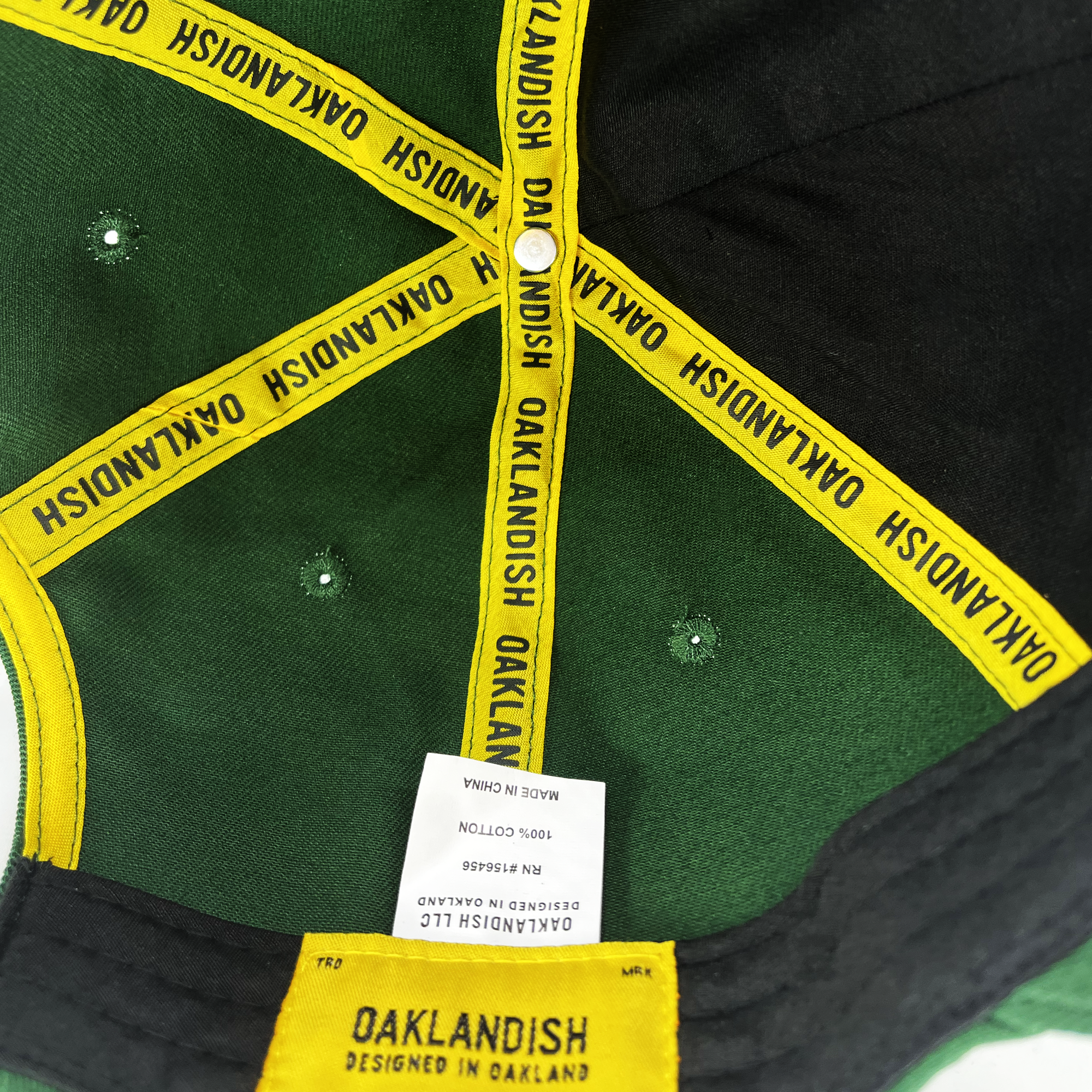 The inside view of the Oakland Flag Dad Hat reveals a green and black baseball cap. Yellow ribbons with "OAKLANDISH" text line the seams, reminiscent of the Oakland flag's vibrance. A white tag in the center reads "Made in China / 100% Cotton / RN#104866." The rear label says "OAKLANDISH - DESIGNED IN OAKLAND." The grom