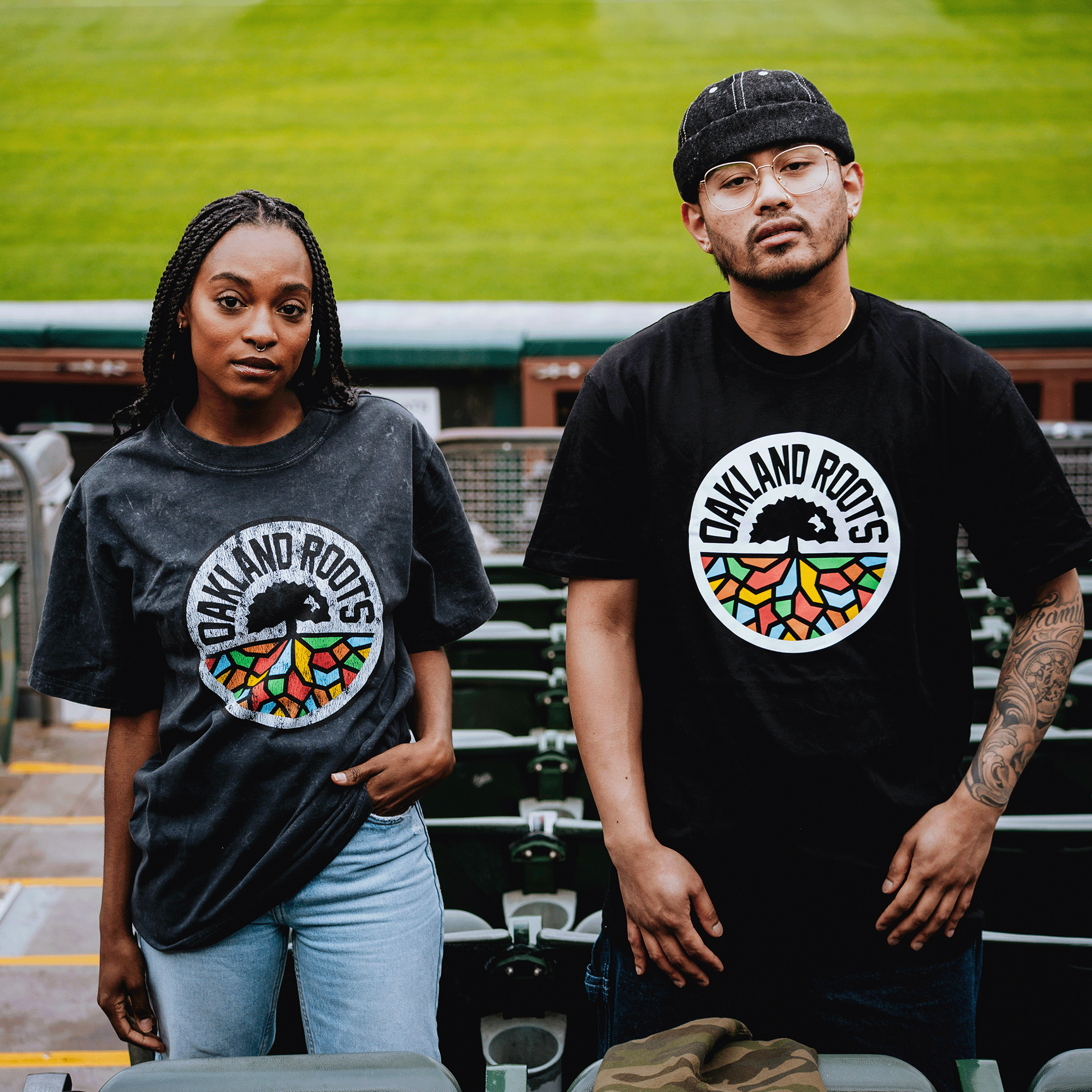 Two people in stadium seating wear black Oakland Roots SC Classic Tees by Oakland Roots SC, each with a colorful tree logo. The woman on the left has long braids; the man on the right wears glasses, a cap, and reveals his tattooed arm.