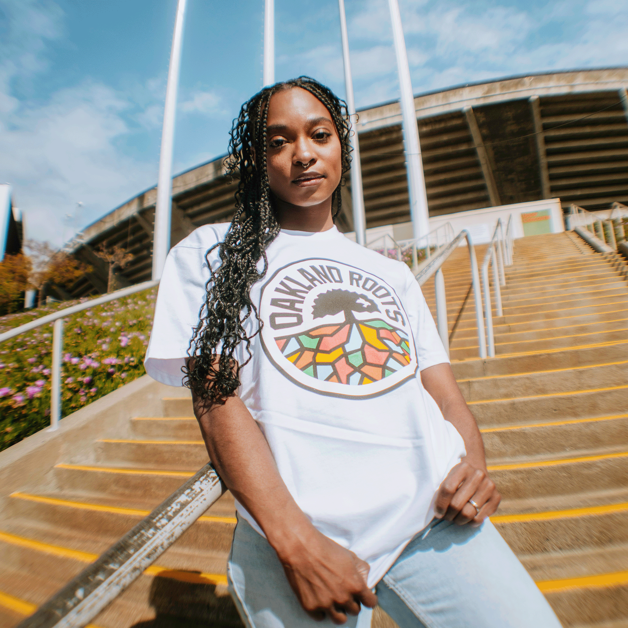 A woman with long braided hair stands confidently on outdoor steps, wearing the Oakland Roots SC Classic Tee by Oakland Roots SC. Behind her are yellow-striped stairs, metal railings, flowering plants, and a stadium with flagpoles.