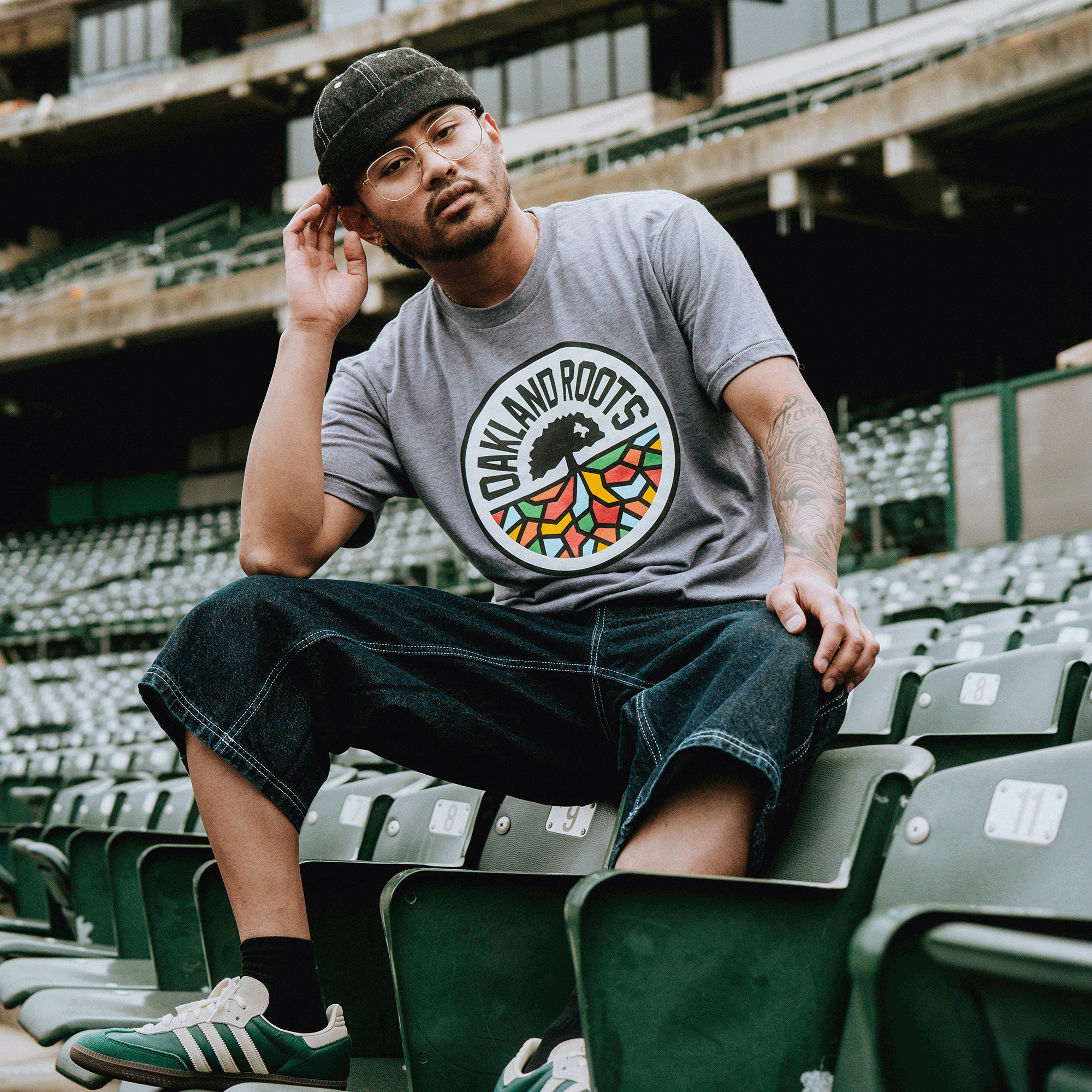 Wearing glasses, a dark knit cap, denim shorts, and sneakers, a man in a gray Oakland Roots SC Classic Tee by Oakland Roots SC sits confidently on stadium seats, touching his cap and reflecting the spirited energy of the team.
