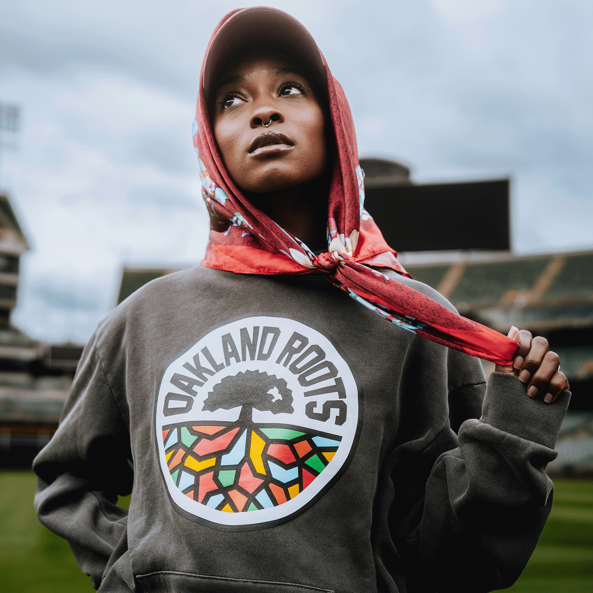 A woman in a red scarf and gray Oakland Roots SC Heavy Hoodie stands in a stadium, gazing upward with confidence. The hoodie, by Oakland Roots SC, displays a tree logo with colorful geometric roots.
