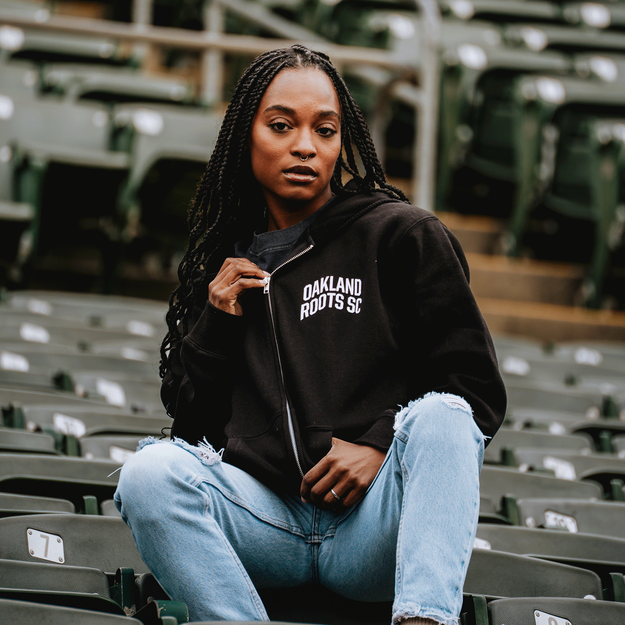 A woman with long braids sits on green stadium seats, confidently looking at the camera while wearing the Oakland Roots SC Classic Zip Hoodie by Oakland Roots SC and light blue ripped jeans, holding the zipper of her hoodie.