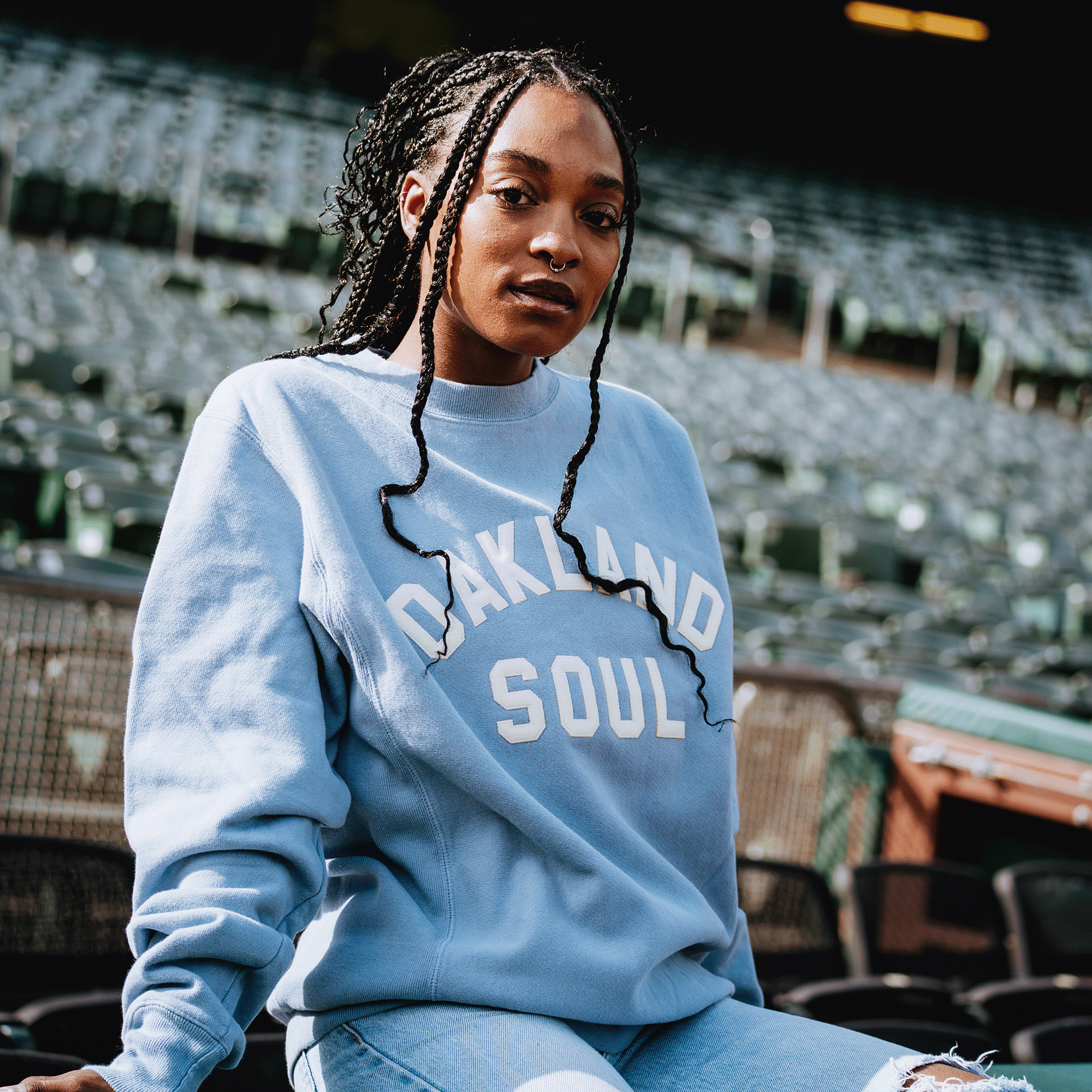 A young woman with braided hair sits in empty stadium seats, wearing the Champion X Oakland Soul Wordmark Crew by Oakland Soul—proudly representing women’s soccer in the USL W-League. Sunlight highlights her confident gaze and the rows behind her.