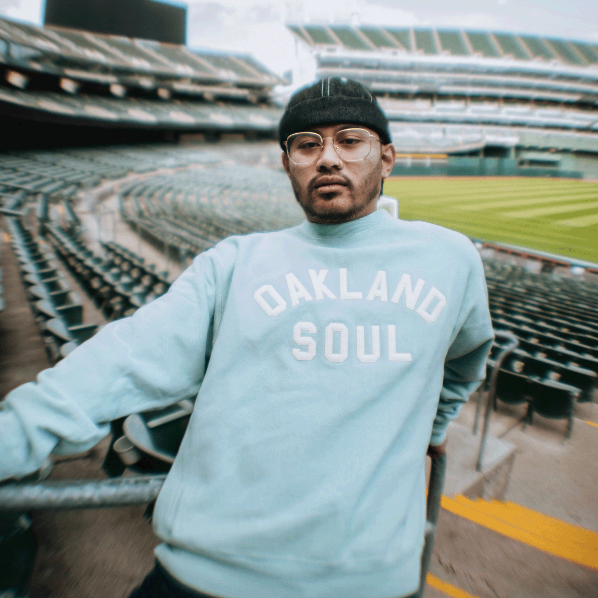 A person in glasses, a black beanie, and the Champion X Oakland Soul Wordmark Crew by Oakland Soul stands by green seats in an empty stadium and baseball field, representing women's soccer and social good with a relaxed look at the camera.