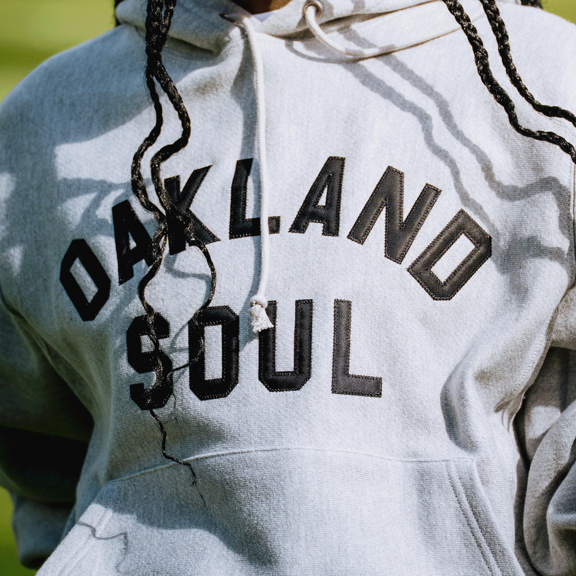 Wearing the Champion X Oakland Soul Wordmark Hoodie—light gray with bold black lettering—a person with long braids stands against a softly blurred, sunlit green backdrop. Oakland Soul is the city’s USL W-League women’s soccer team.