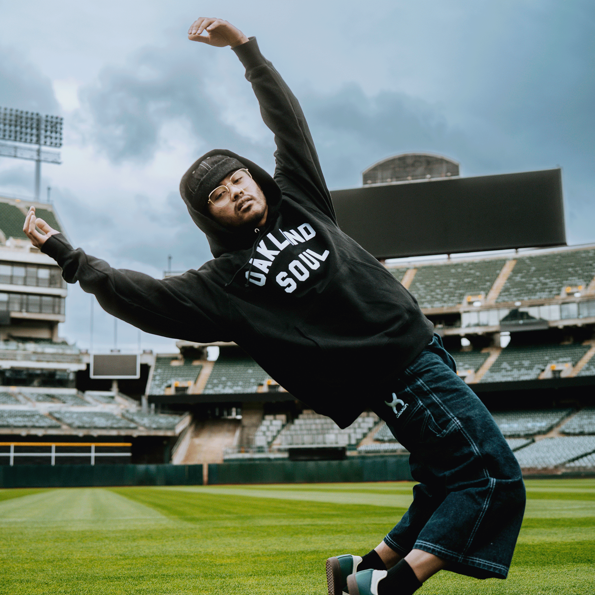 Wearing a Champion X Oakland Soul Wordmark Hoodie, glasses, and a cap, a person strikes a dance pose on the field under cloudy skies. Empty stadium seats and a scoreboard highlight Oakland Soul’s USL W-League women’s soccer spirit.