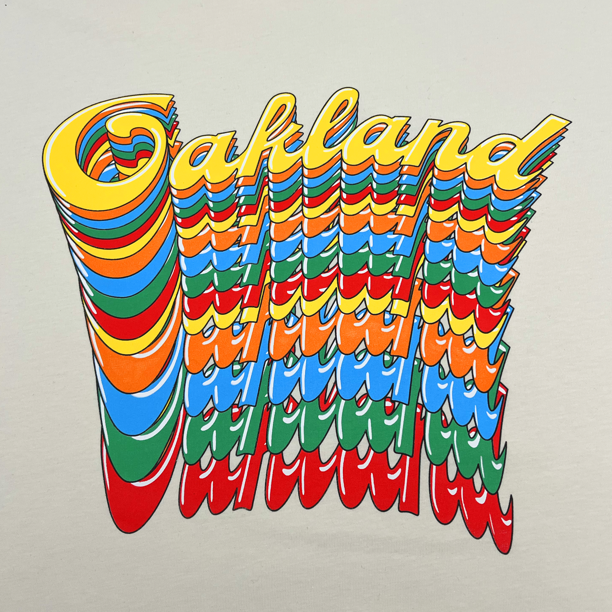 Detail close-up of Oakland wordmark in script on repeat in rainbow color on the front chest of a natural cotton color t-shirt.