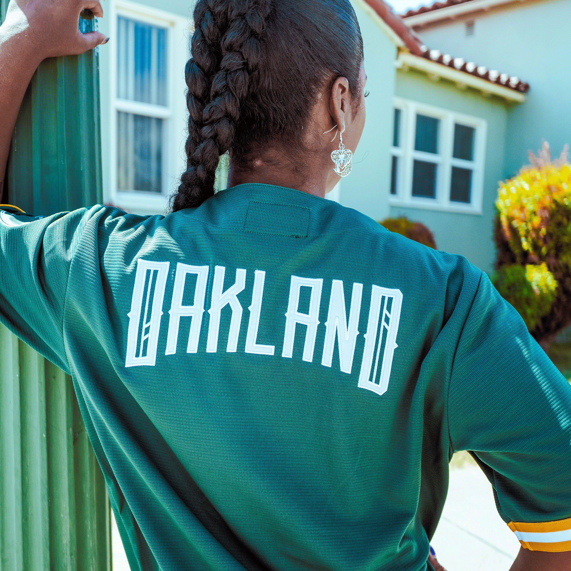 A person with braided hair and heart-shaped earrings wears the Oakland Ballers Button Up Jersey by Oakland Ballers, featuring “OAKLAND” in large white letters on the back, standing outdoors near a green pole with a light blue house and garden behind.