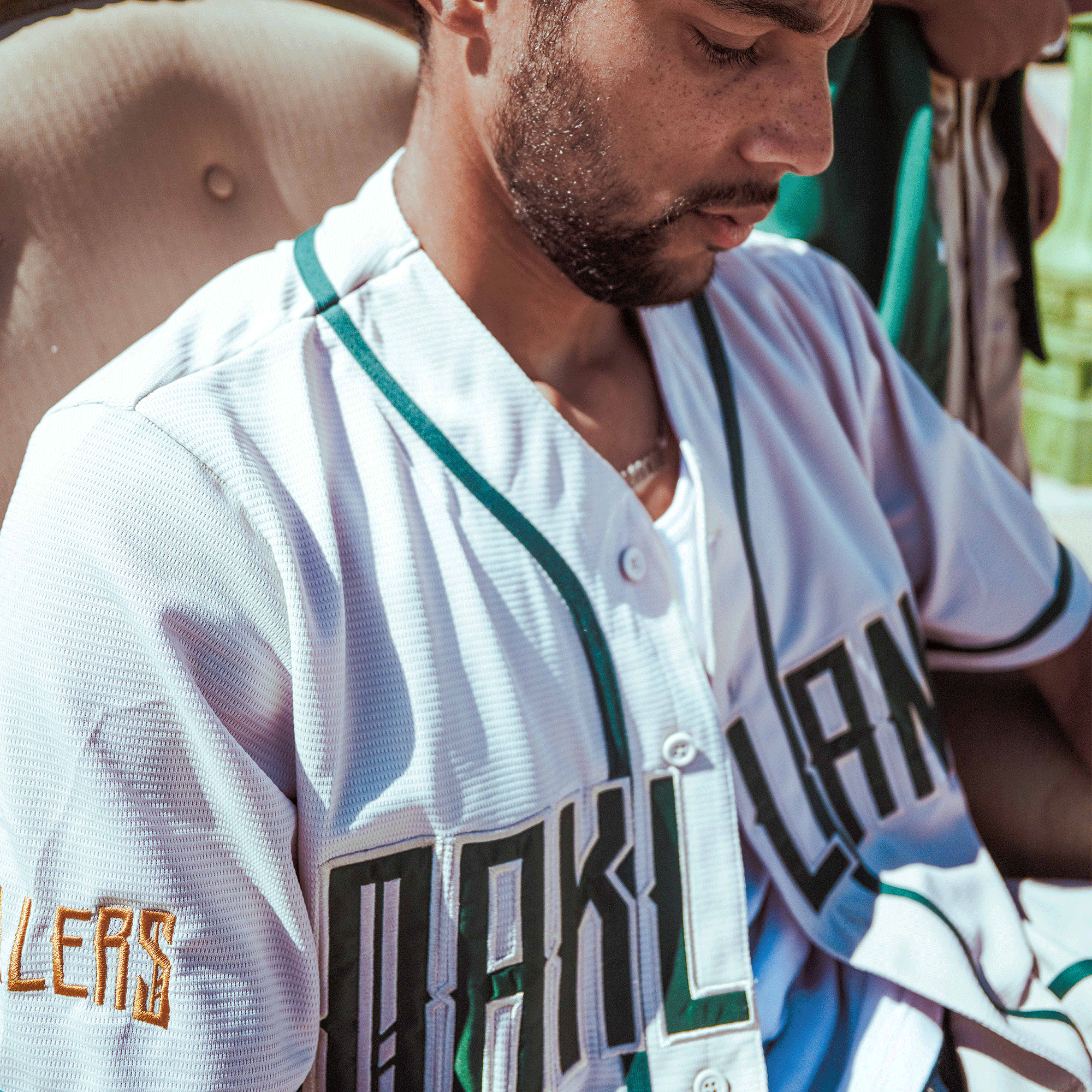 A man in an Oakland Ballers Button Up Jersey, featuring white fabric with green trim and yellow accents, sits looking down. The word “HUSTLERS” appears on his sleeve as sunlight casts shadows across his focused face.