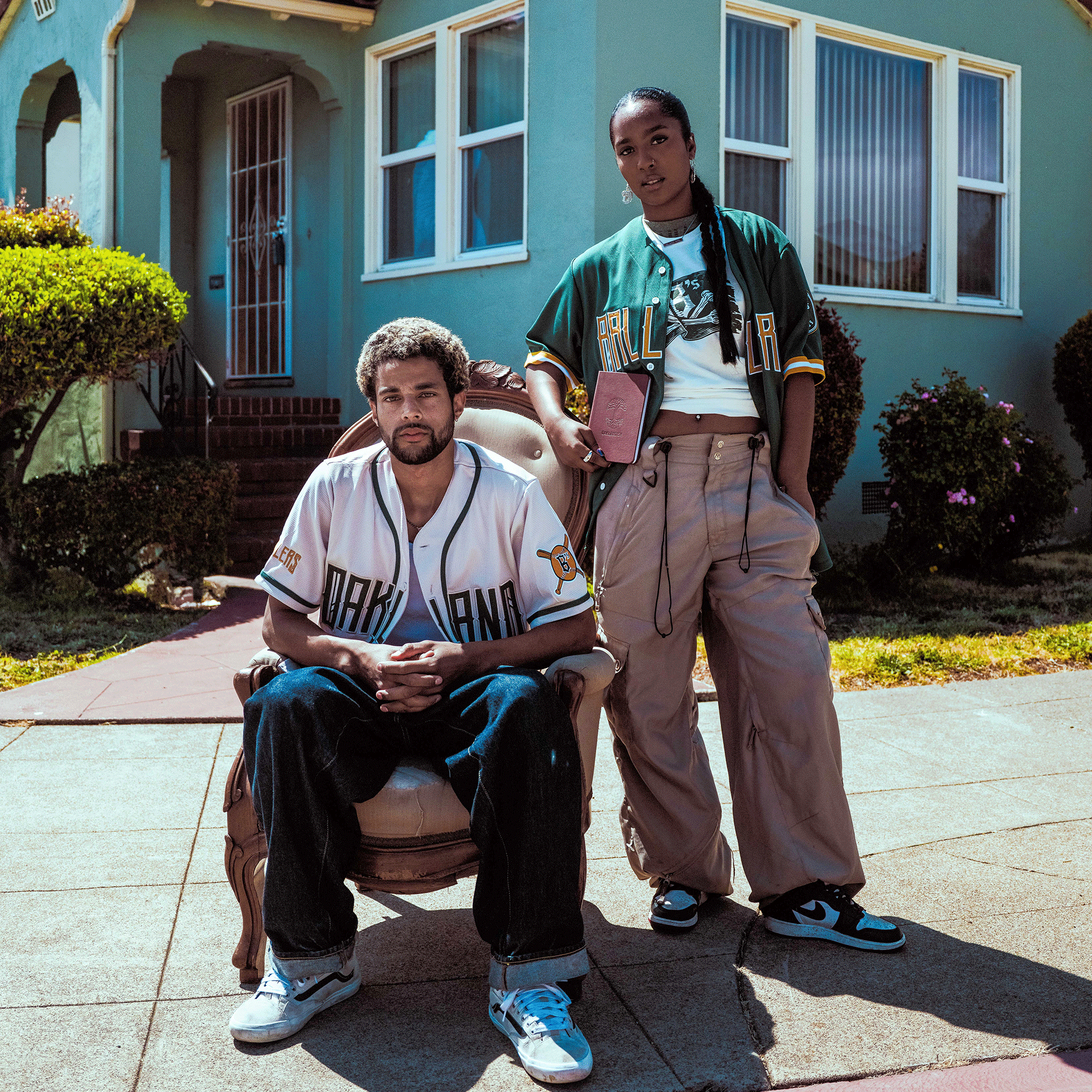 Two young adults pose outside a turquoise house. One sits in an Oakland Ballers Button Up Jersey and jeans, hands clasped; the other stands beside in a green jersey, tan pants, and sneakers, holding a book. Sunlight and shrubs frame the scene.