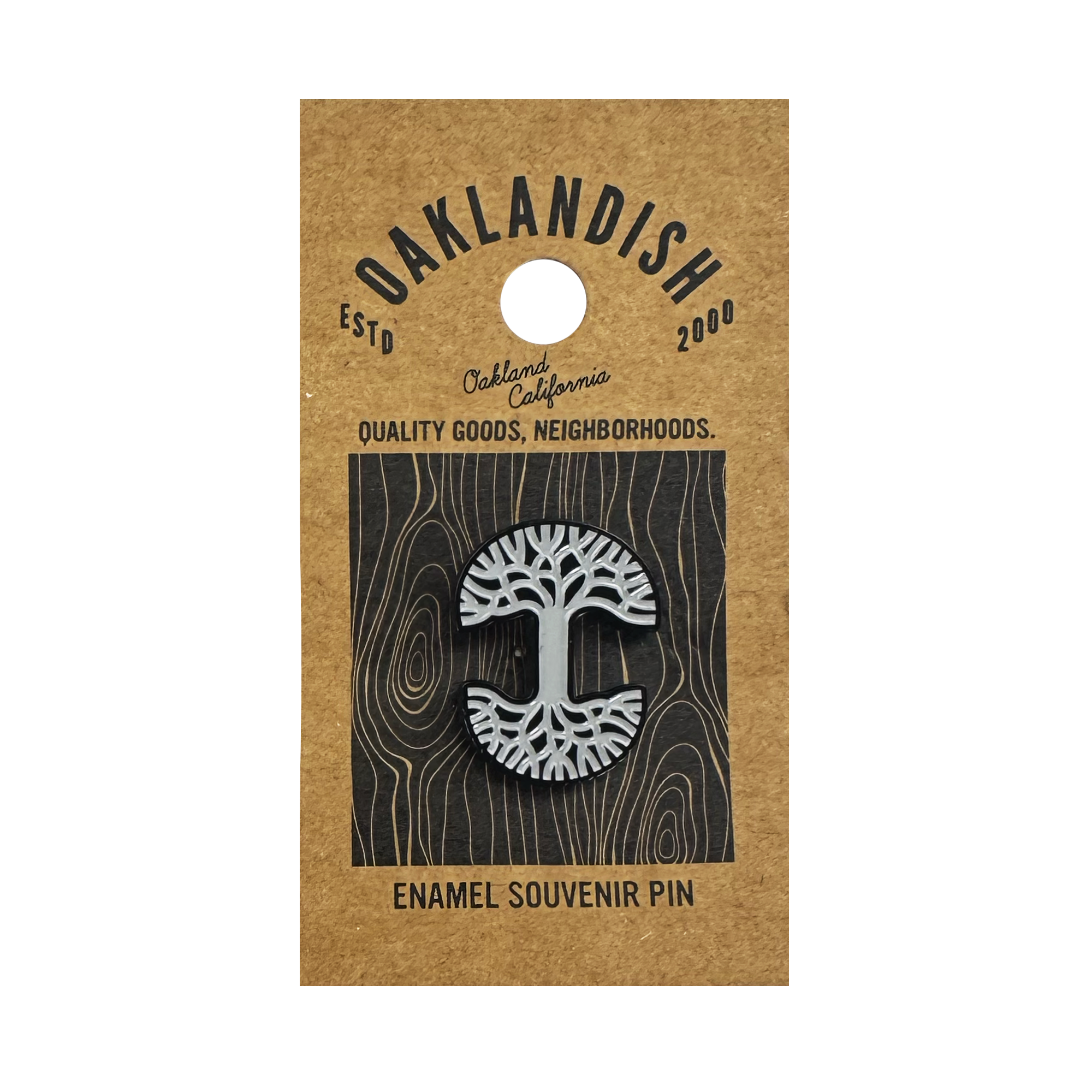 The Oaklandish Logo Enamel Pin by Oaklandish features a black and white enamel abstract tree on a brown kraft card reading "OAKLANDISH ESTD 2000, Oakland California, Quality Goods, Neighborhoods" with a grey woodgrain background.